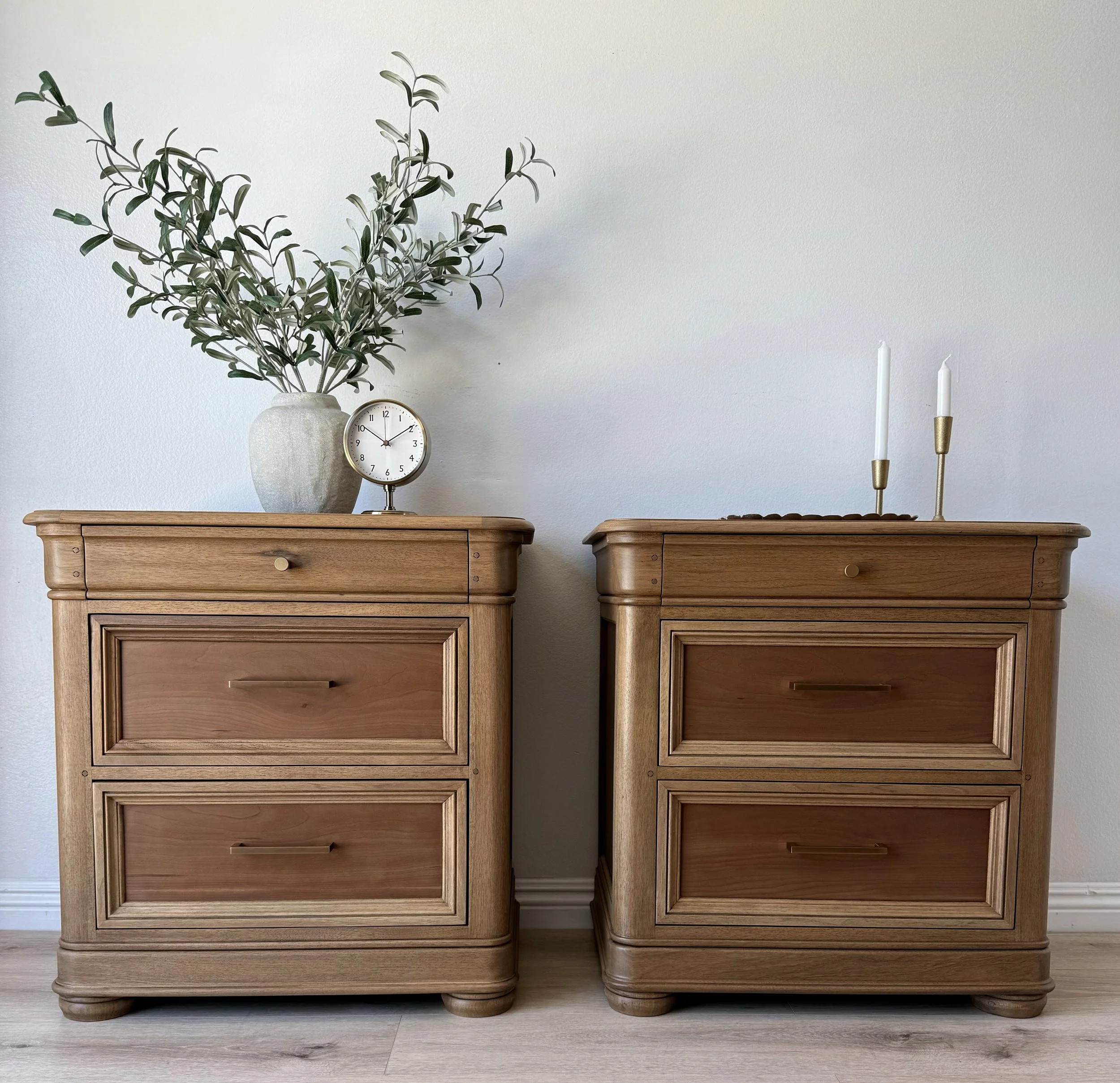 Two wooden nightstands with drawers, decorated with a large potted plant and a clock on the left nightstand, and two white candles on brass holders on the right nightstand, against a white wall.