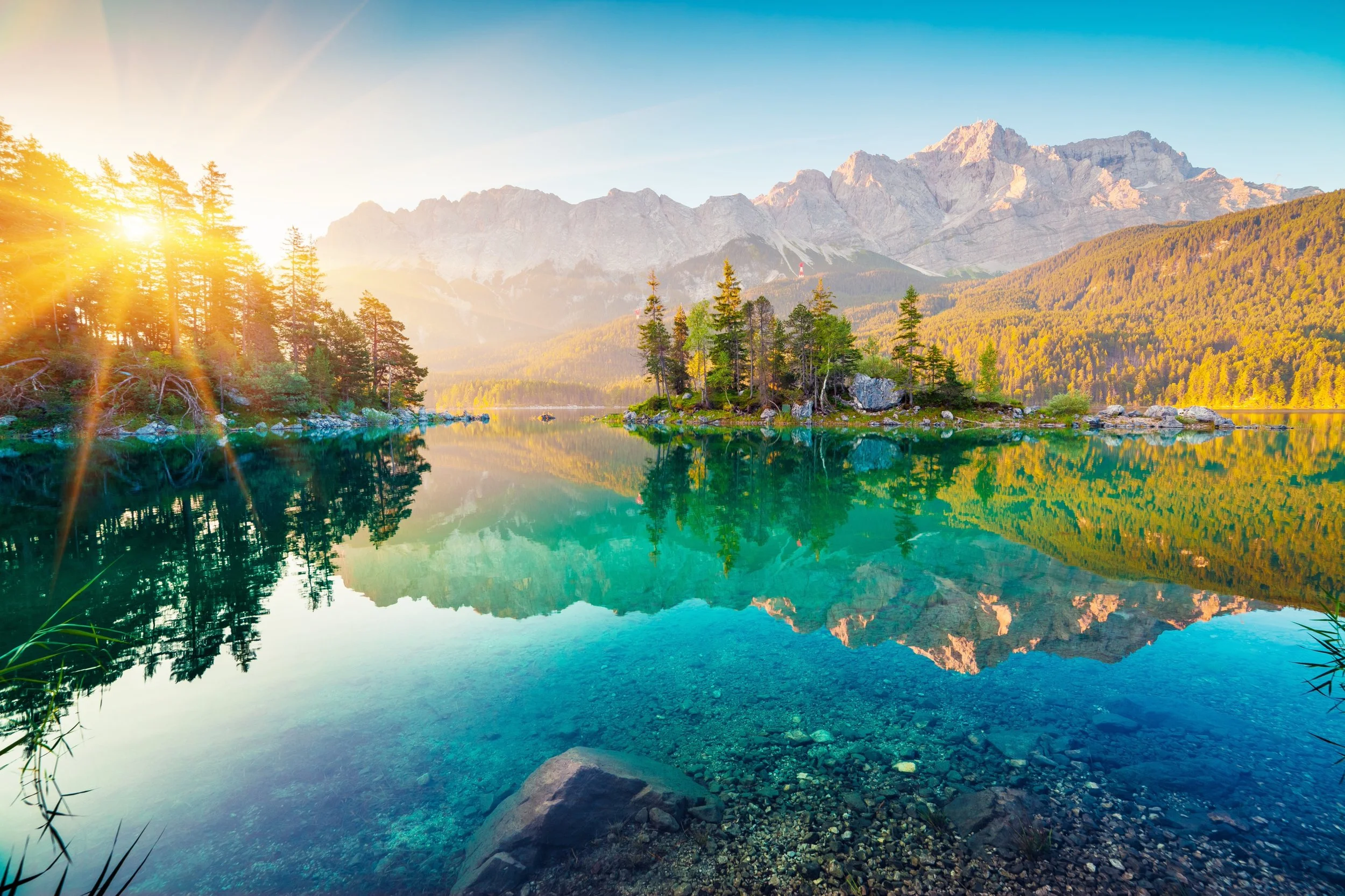Sunrise over a clear mountain lake with green trees, rocky shoreline, and mountains in the background.