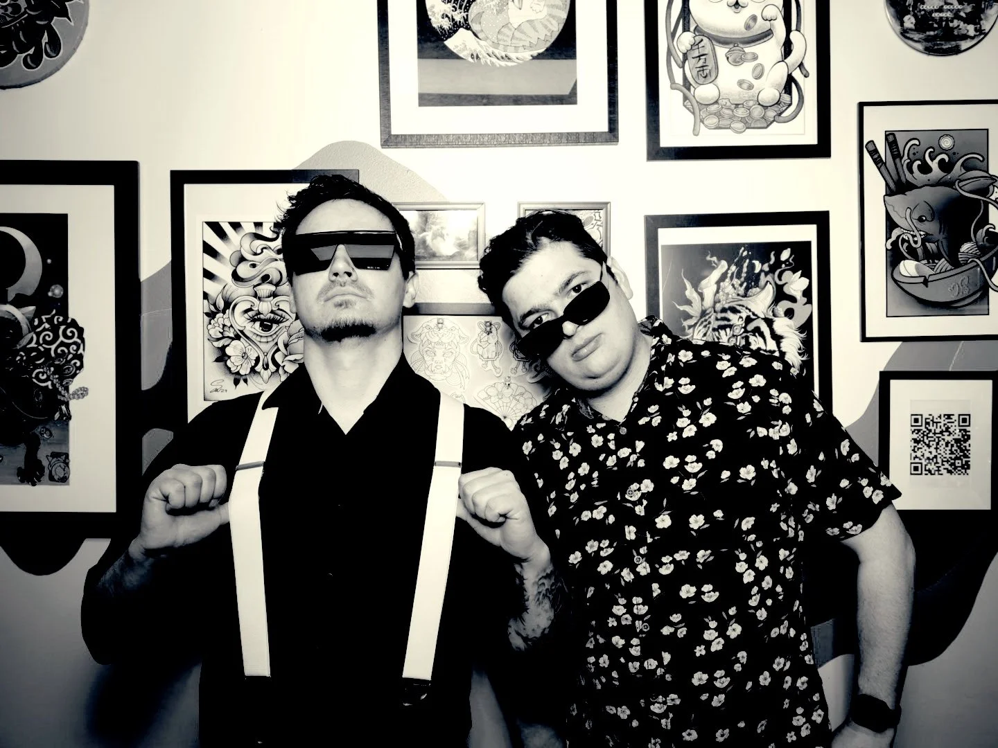 Two young men wearing sunglasses pose in front of a wall decorated with various framed artwork and illustrations. The man on the left is dressed in black with suspenders, and the man on the right is wearing a floral shirt. Both are making a fist gesture with their right hands.