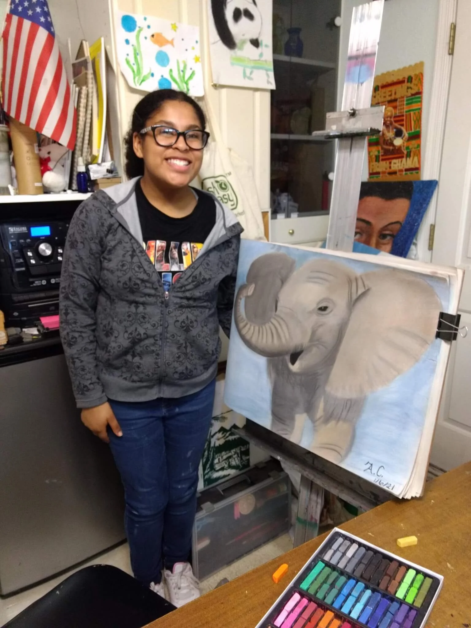 A young woman smiling, standing in an art studio with a painting of an elephant on an easel. The studio has colorful artwork hanging on the wall, an American flag, and various art supplies visible.