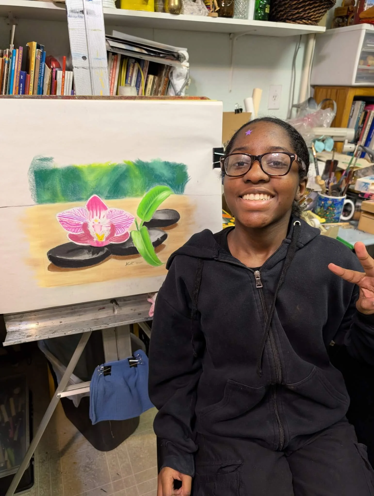 A young girl wearing glasses and a black hoodie sitting in front of an art painting in a cluttered art studio, smiling and making a peace sign.