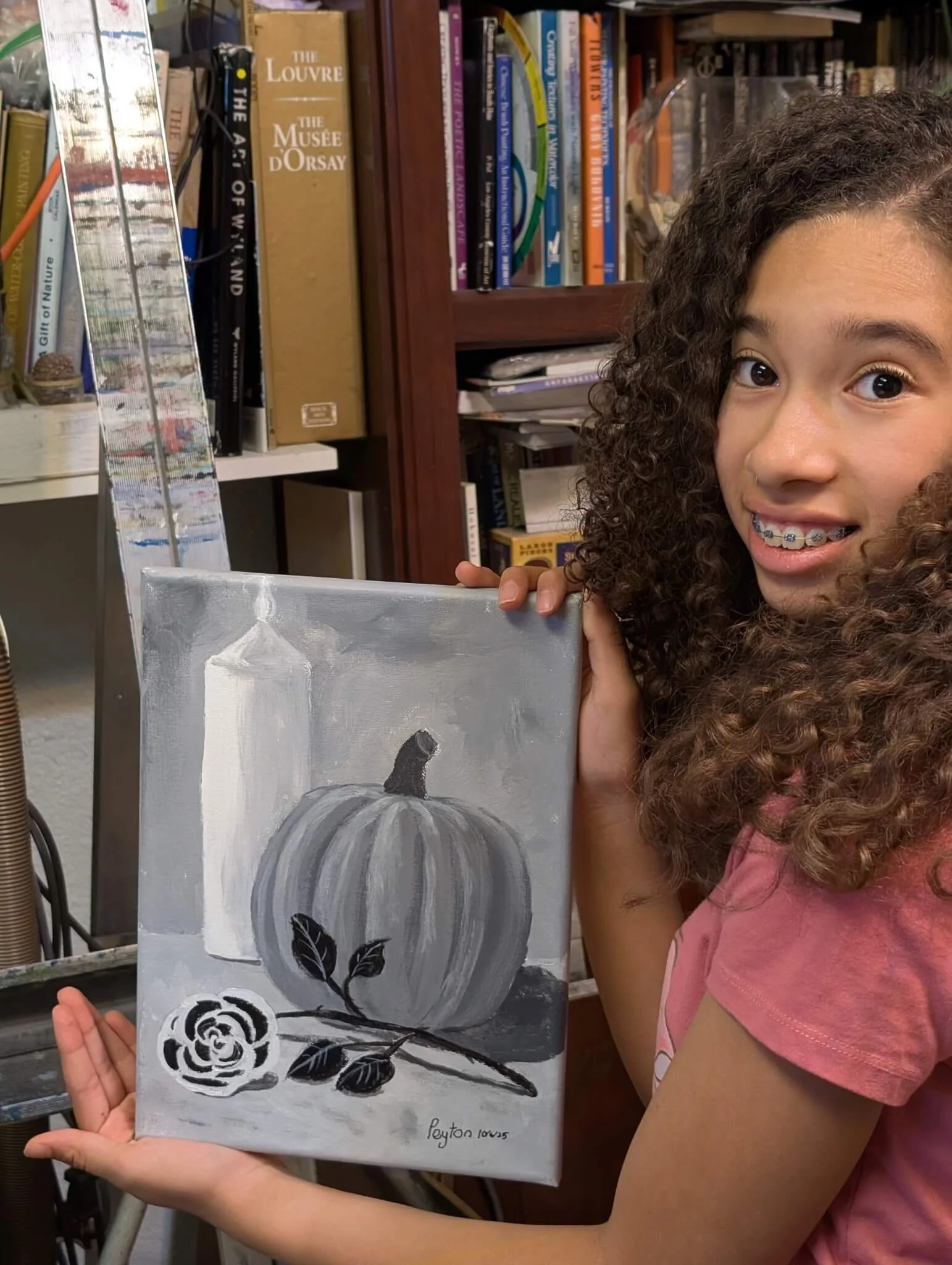 A young girl with curly hair and braces holding a small painting of a pumpkin, a rose, and a bottle in grayscale.