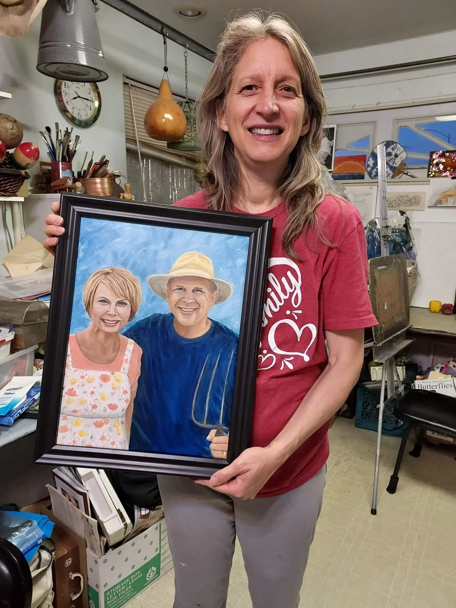 A woman with long blonde hair smiling and holding a framed portrait of an elderly couple, the woman in the portrait wearing a floral apron and the man wearing a wide-brimmed hat and holding a pitchfork, standing in a workshop or studio.