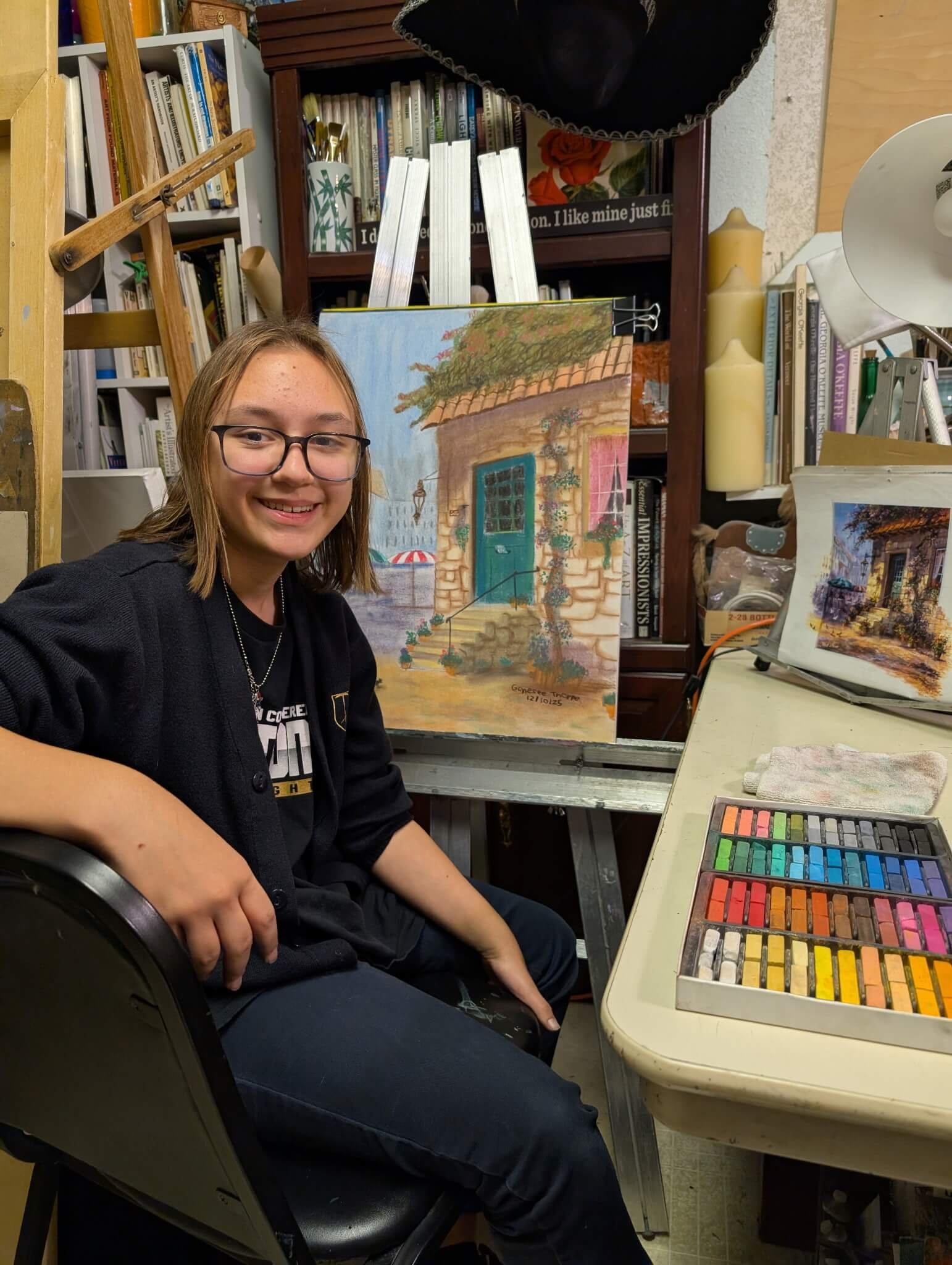 Young girl with glasses sitting at an art station, smiling, with an unfinished painting of a door and plants on an easel behind her, art supplies including pastel chalks on the table, and shelves filled with books and decorative items in the backgrou