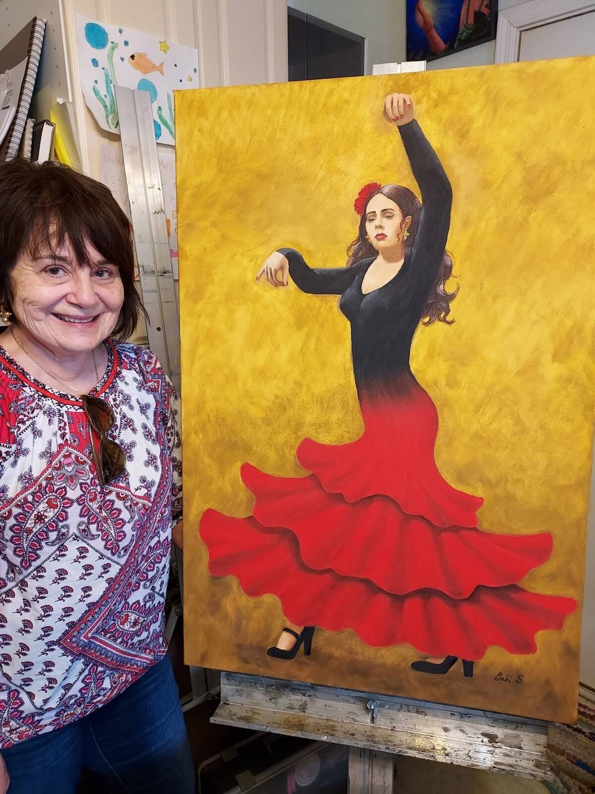 A woman stands beside a painting of a flamenco dancer in a black top and red ruffled skirt, with a yellow background. The woman is smiling and wearing a patterned blouse.