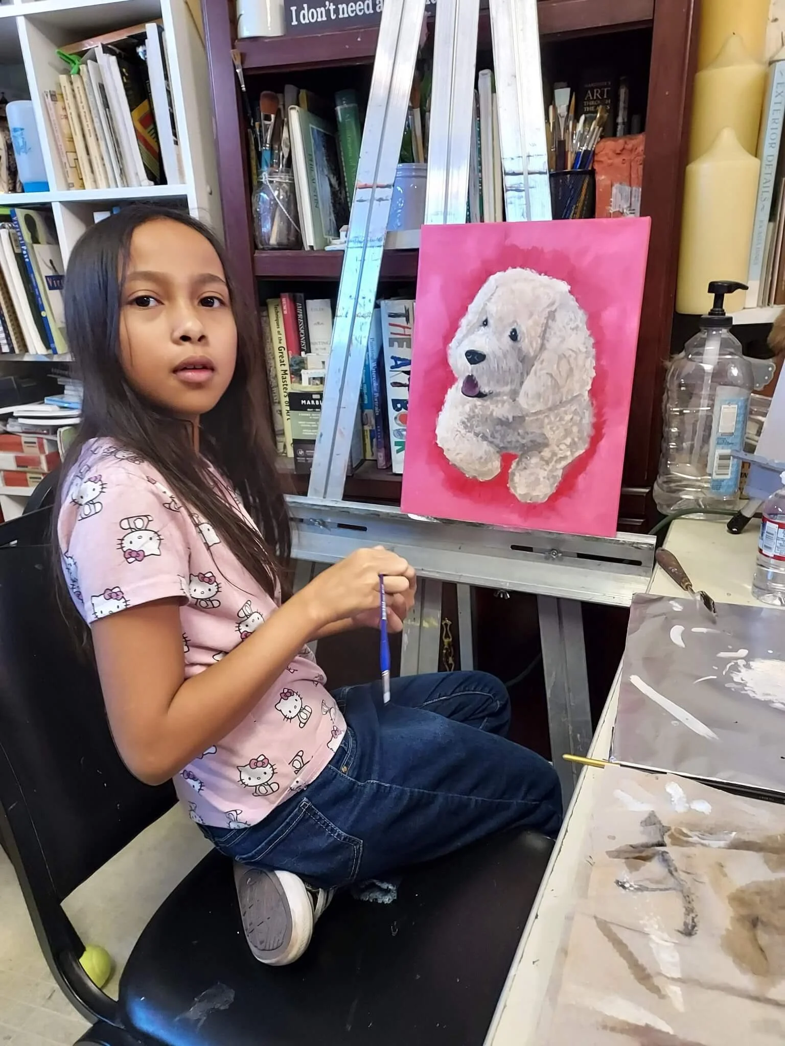 A young girl with long dark hair painting a picture of a fluffy white dog on a pink background in an art studio.