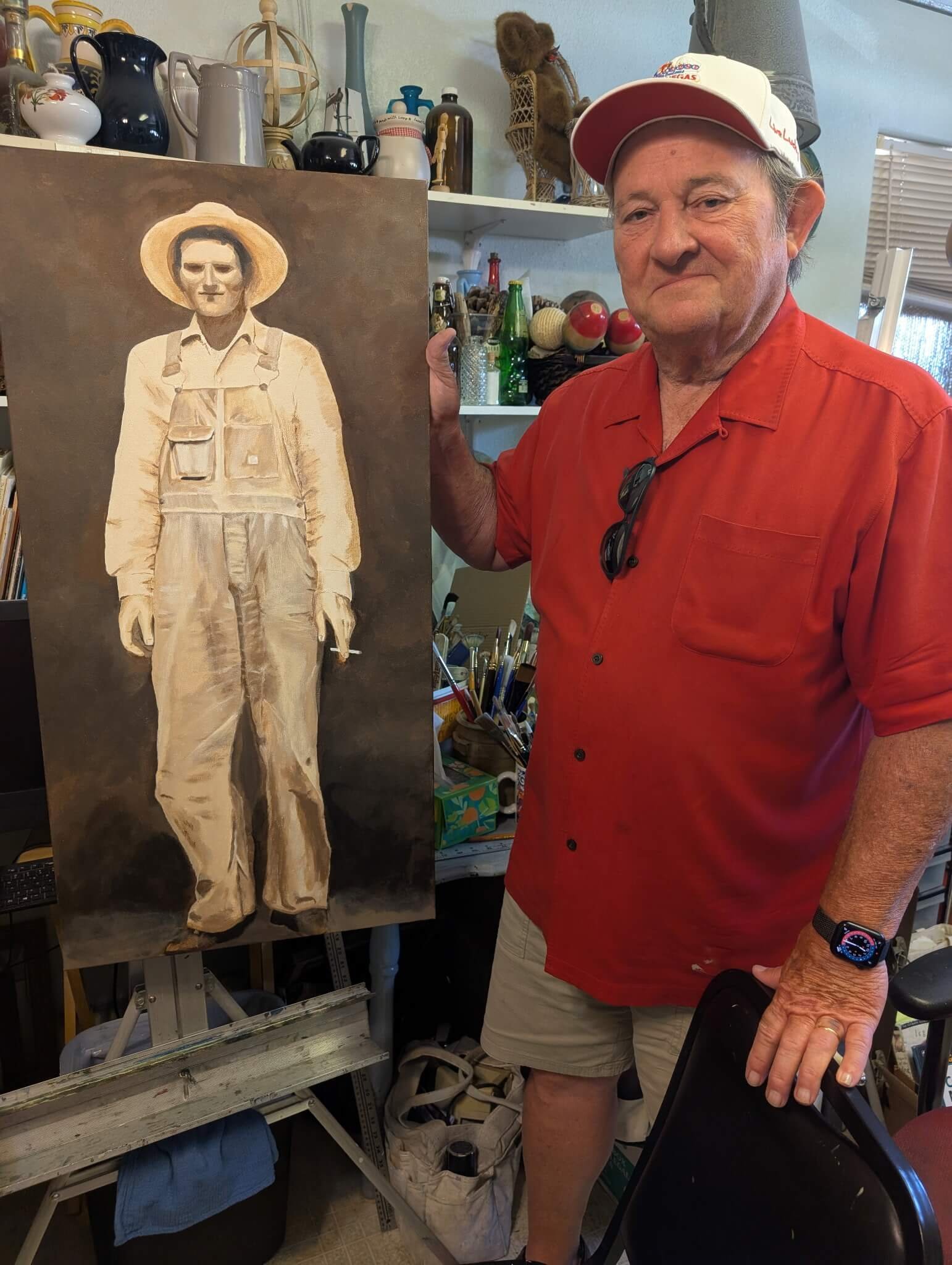 An older man in a red shirt and beige shorts holding a painting of himself in what appears to be a studio or cluttered room with shelves of decorative objects and tools in the background.