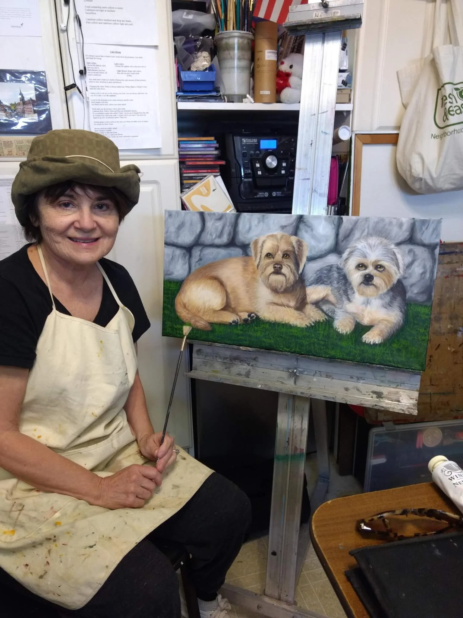 An older woman wearing a paint-stained apron and a hat sitting in her art studio, holding a paintbrush. She is next to a painting of two small dogs lying on grass with a stone wall background.