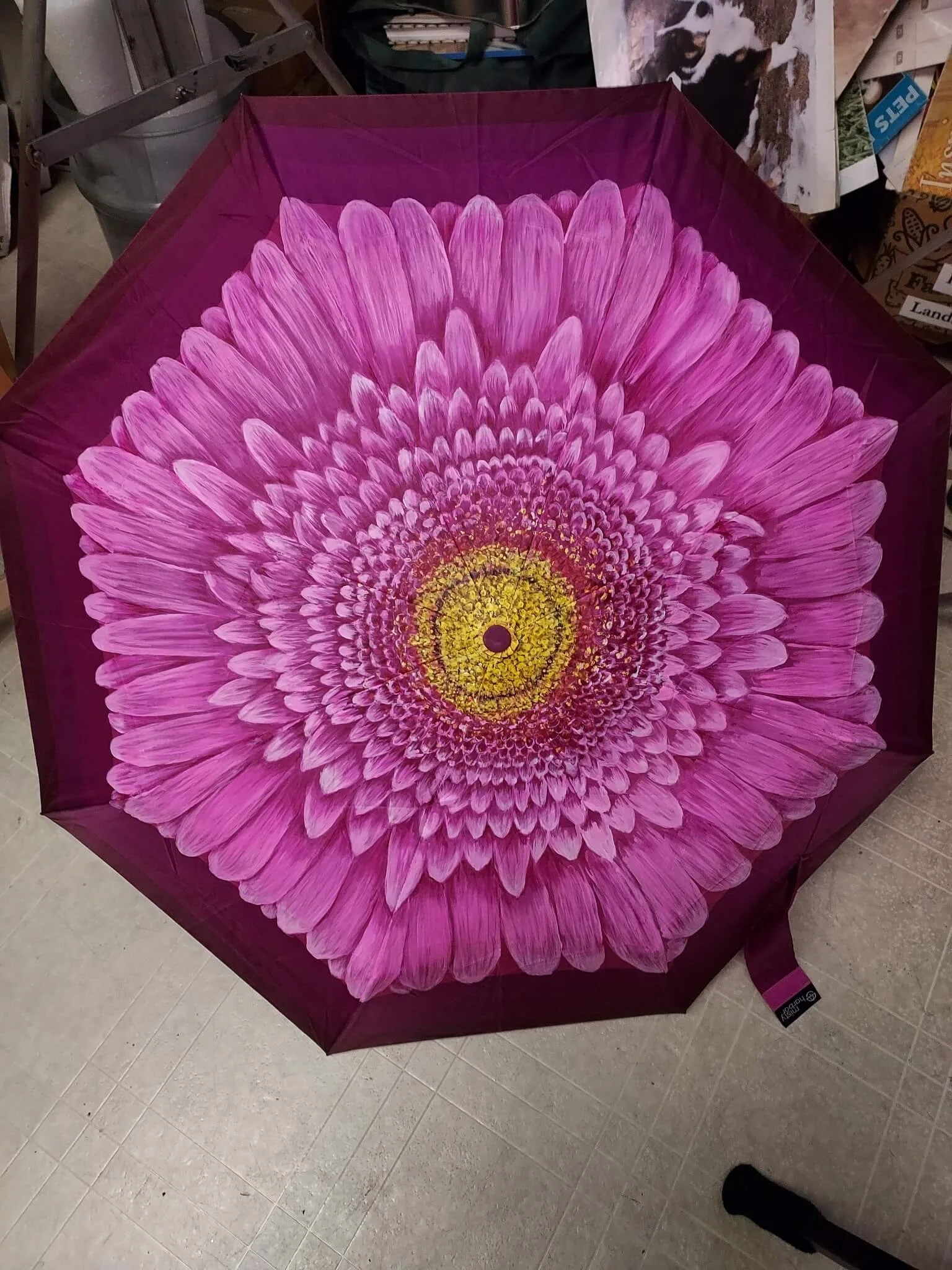 Pink umbrella with floral pattern resembling a large daisy or chrysanthemum with yellow center, placed on a tiled floor in a store.