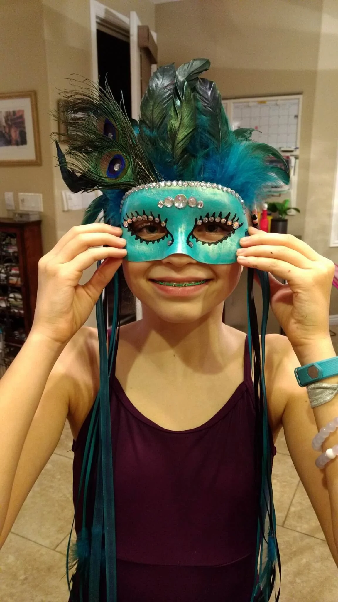 A young girl in a sleeveless maroon dress holds a blue mask decorated with rhinestones, feathers, and peacock plumes, smiling at the camera.