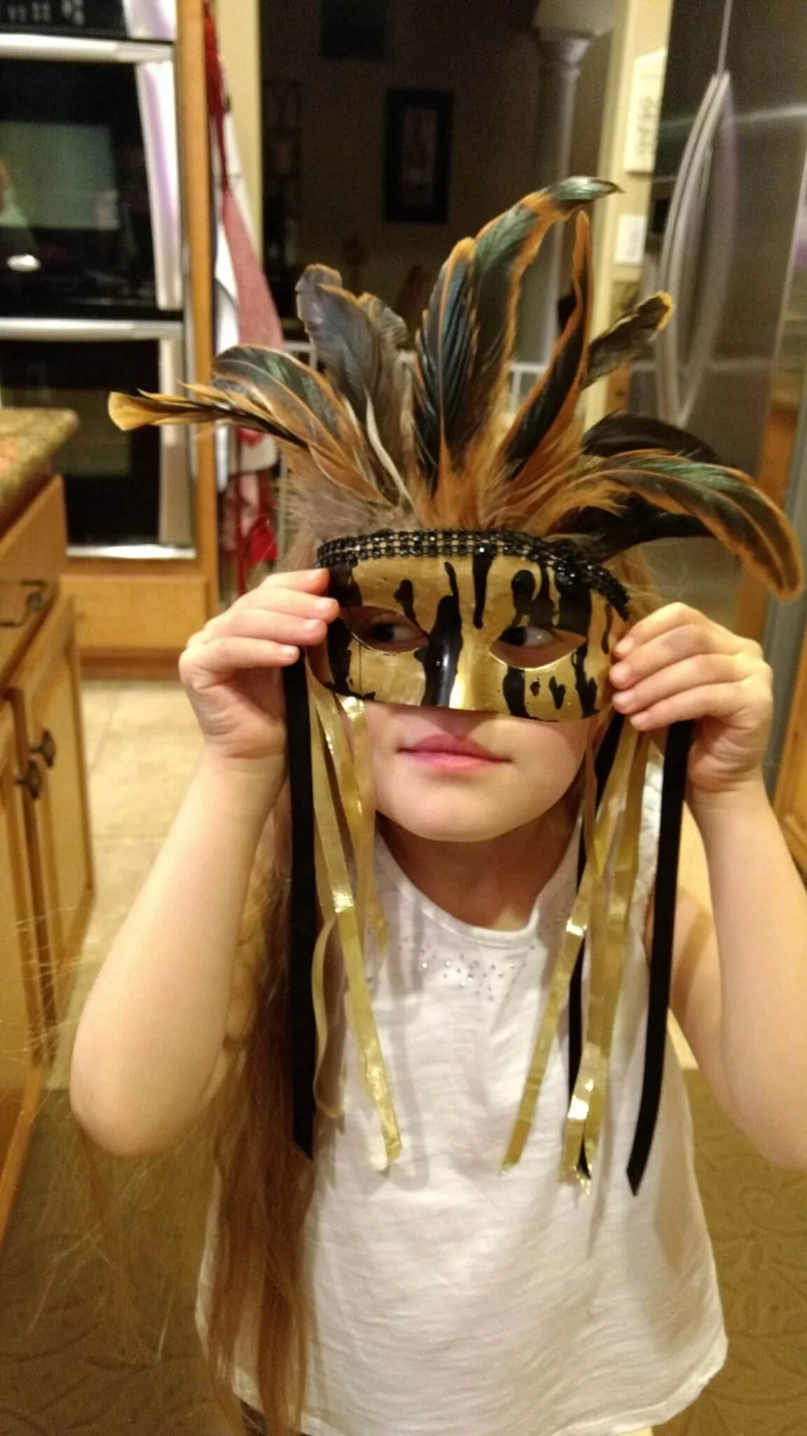 A young girl wearing a gold and black masquerade mask with feathers, ribbons, and decorative details, looking to the side in a kitchen setting.