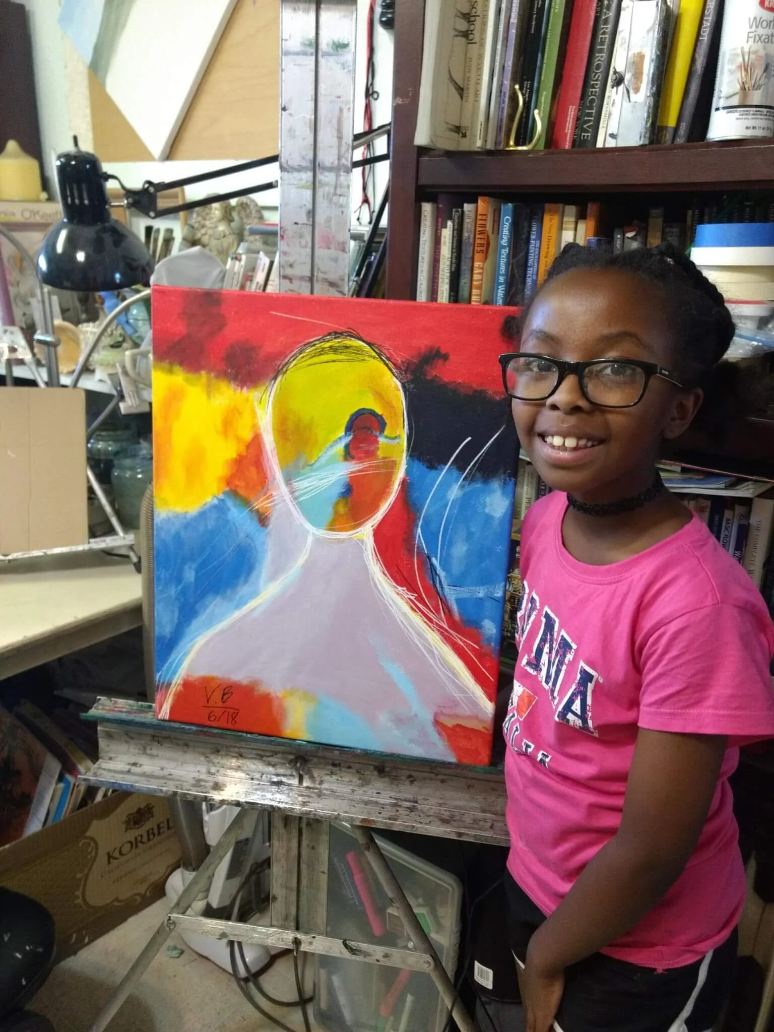 Young girl with glasses and a pink T-shirt smiling next to an abstract colorful painting on an easel.