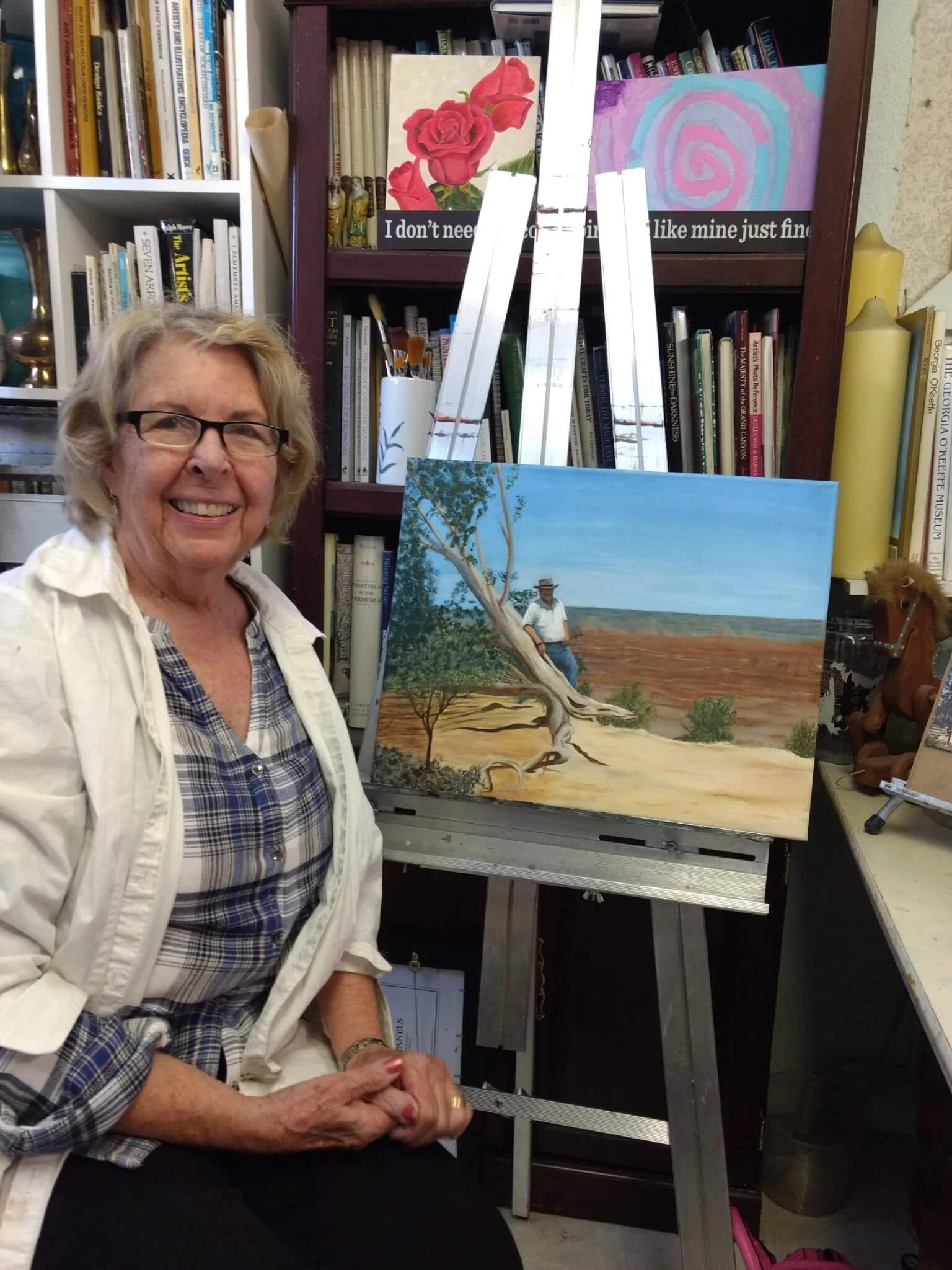 An elderly woman with glasses and short, curly gray hair sitting in front of an easel with a landscape painting of a tree and a man sitting under it, in a room filled with books and art supplies.
