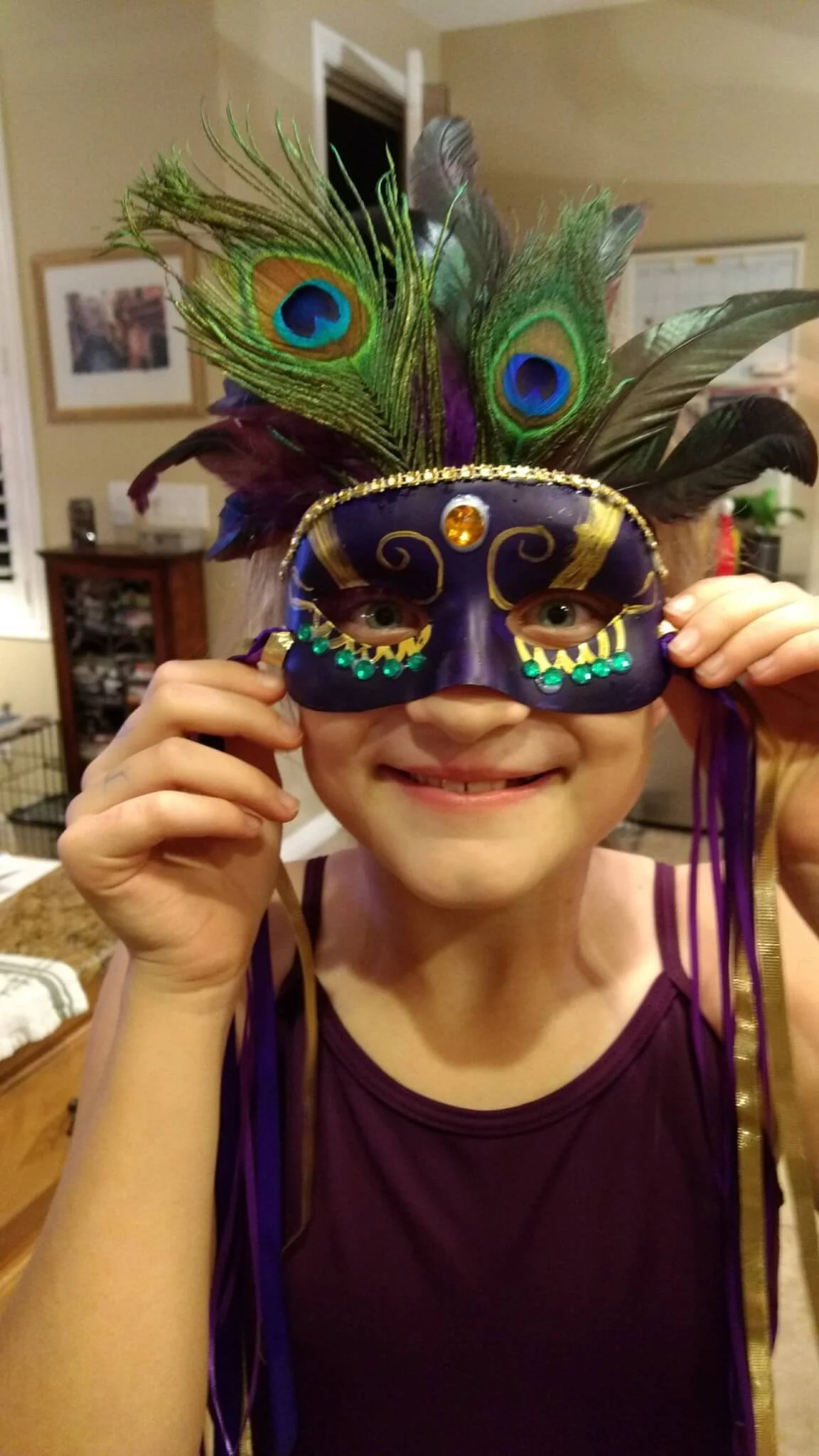 Child wearing a purple and gold costume mask with peacock feathers, smiling while holding the mask in front of their face.