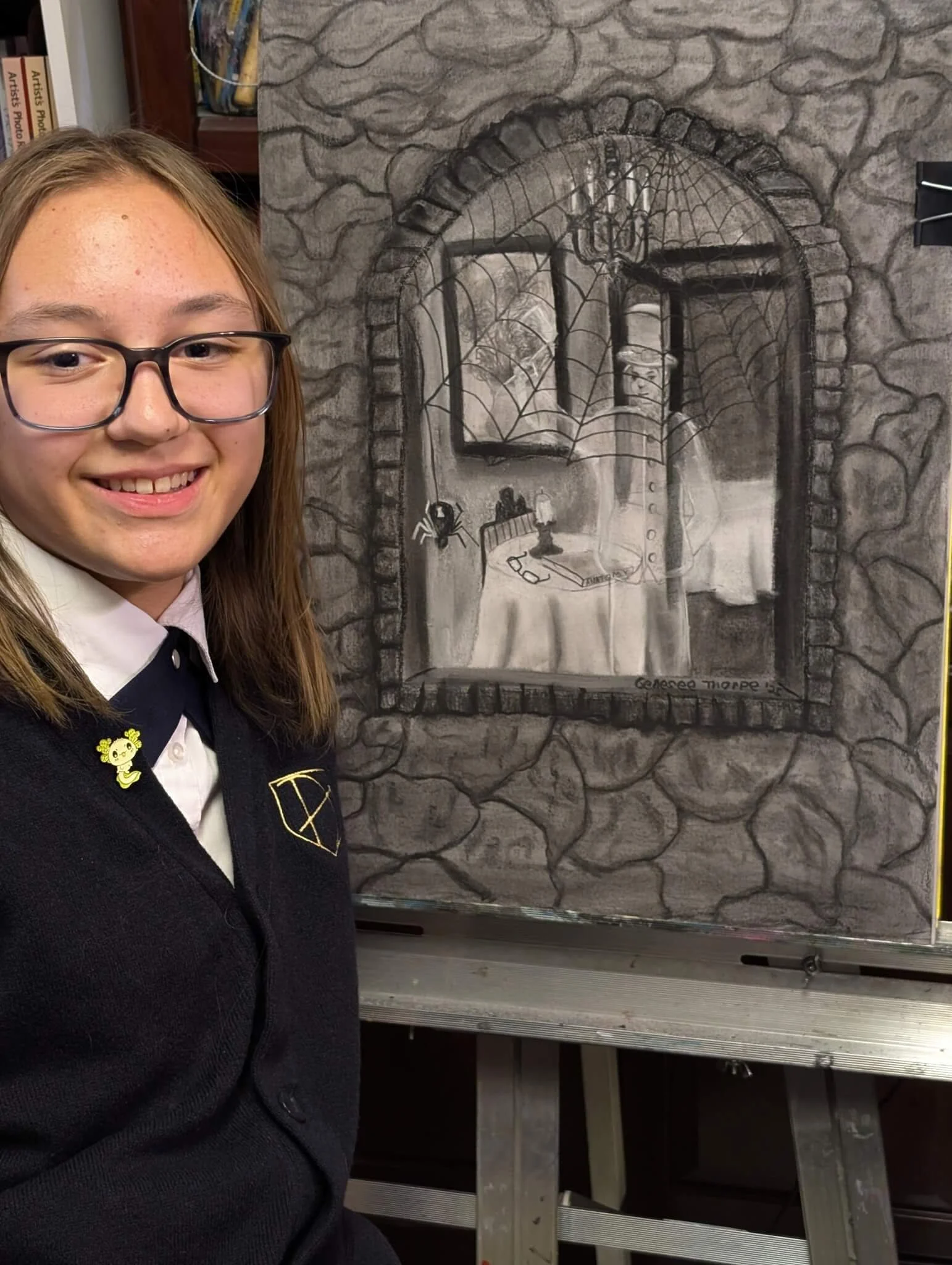 A young girl with glasses and a black school uniform poses in front of a painted mural of a window with a spider hanging from a web. The mural depicts a room with a bed, a lamp, a pair of glasses, and framed pictures.