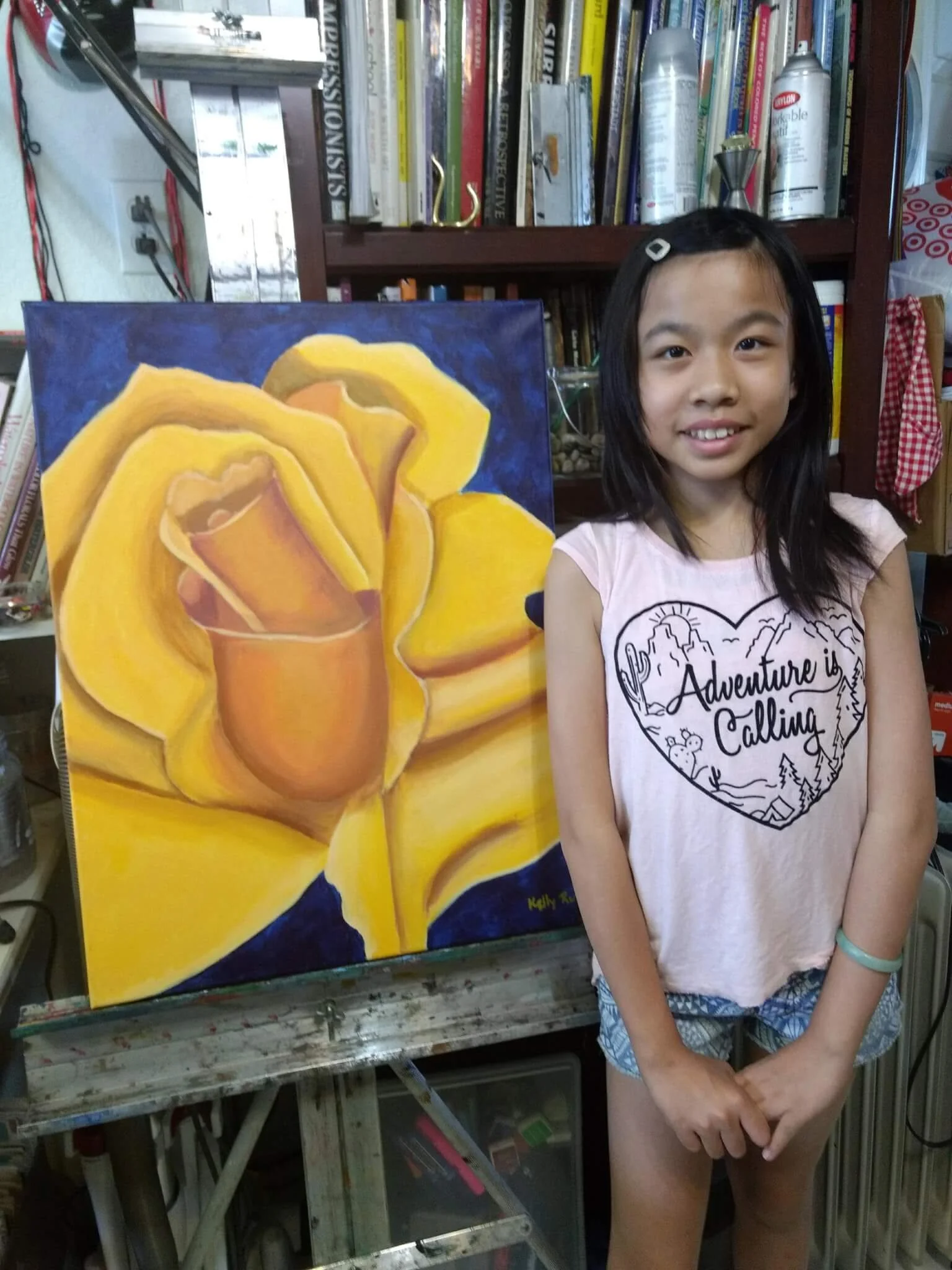 A young girl standing next to a yellow flower painting on an easel, in a room with bookshelves and art supplies.