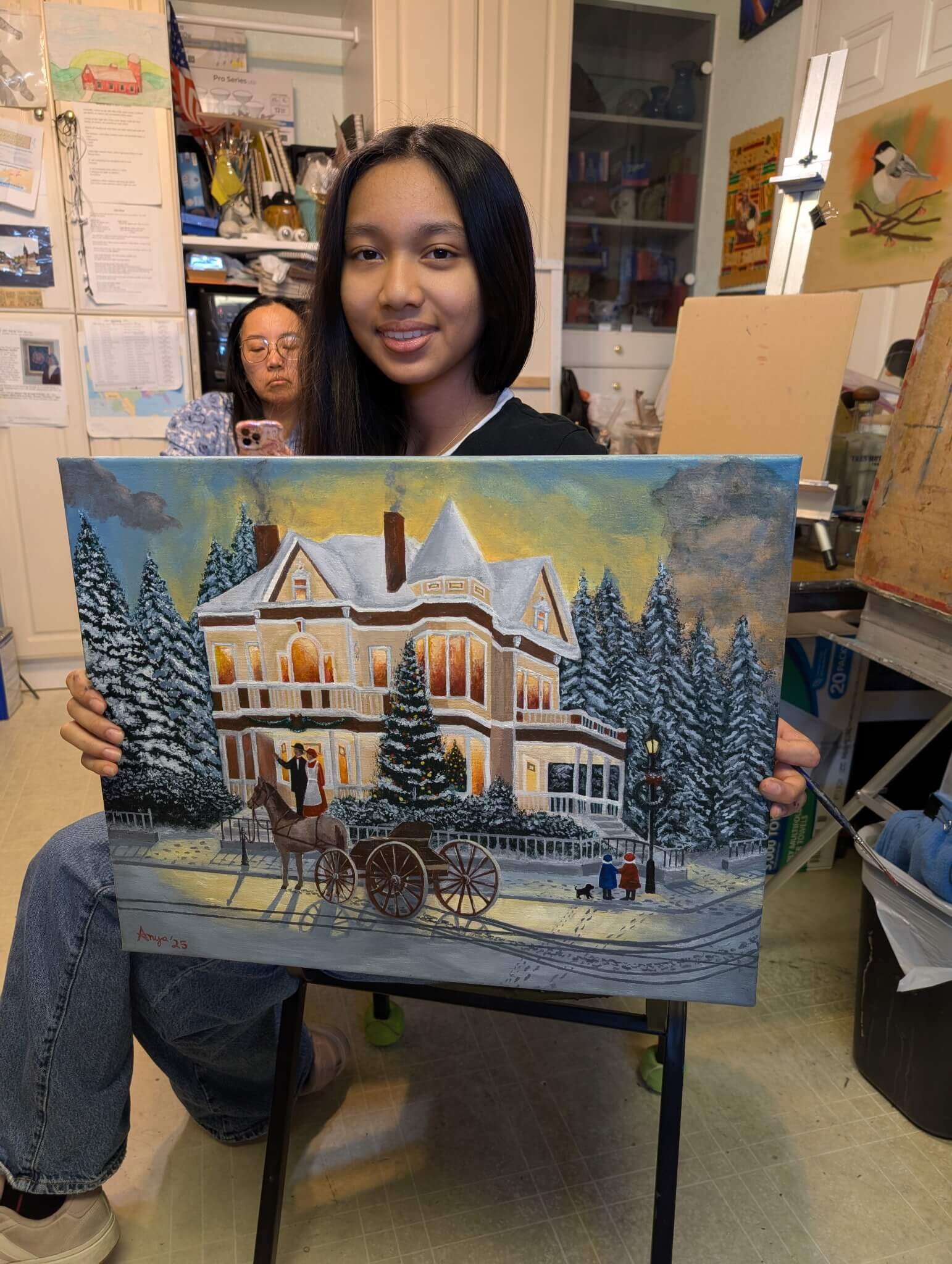 A young woman in an art studio proudly shows her painting of a large, snow-covered Victorian house with a decorated Christmas tree, a horse-drawn carriage, and a couple walking with a dog in the foreground. A woman in glasses is sitting behind her, looking at her phone.