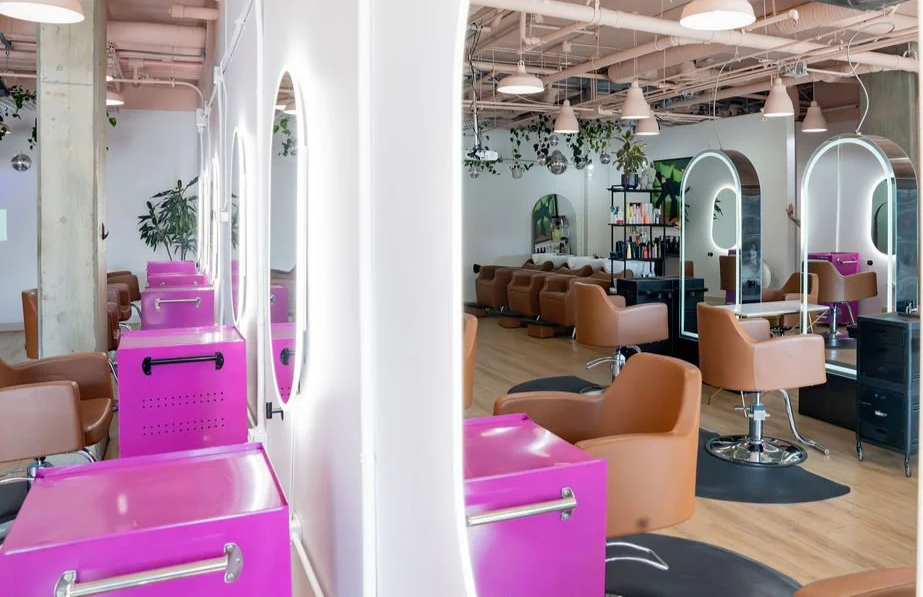 Interior of a modern hair salon with brown chairs, pink styling stations, mirrors, and a shelf with hair products, decorated with green plants and hanging lights.