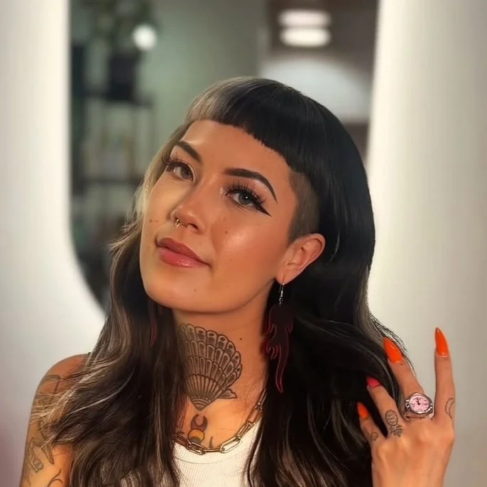 A woman with dark hair and a blunt black bob hairstyle, smiling and pointing at her hair in front of a circular mirror.