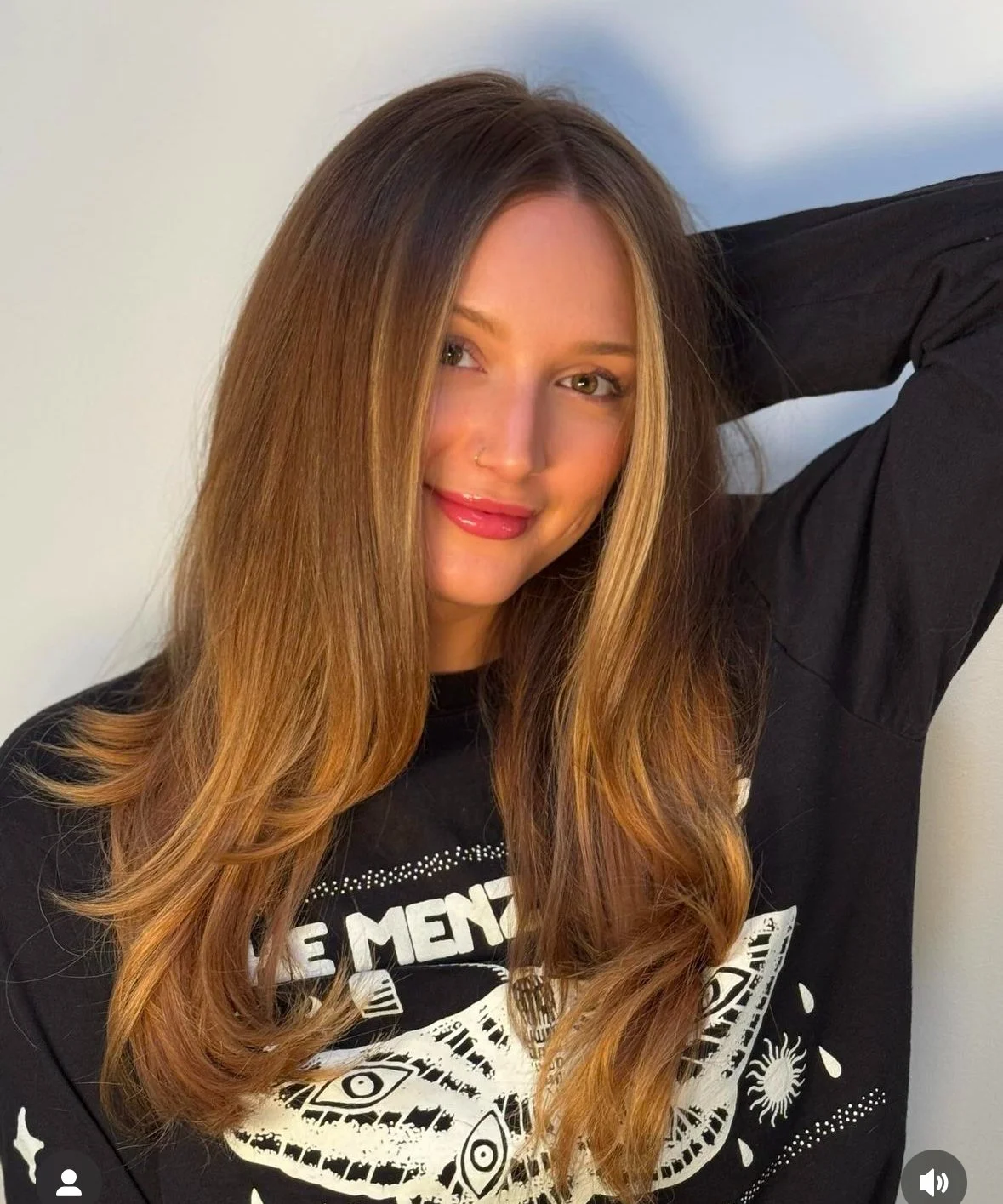 A young woman with long, wavy brown hair, wearing a black shirt with white graphic designs, smiling and posing with her hand on her head, against a plain light-colored background.