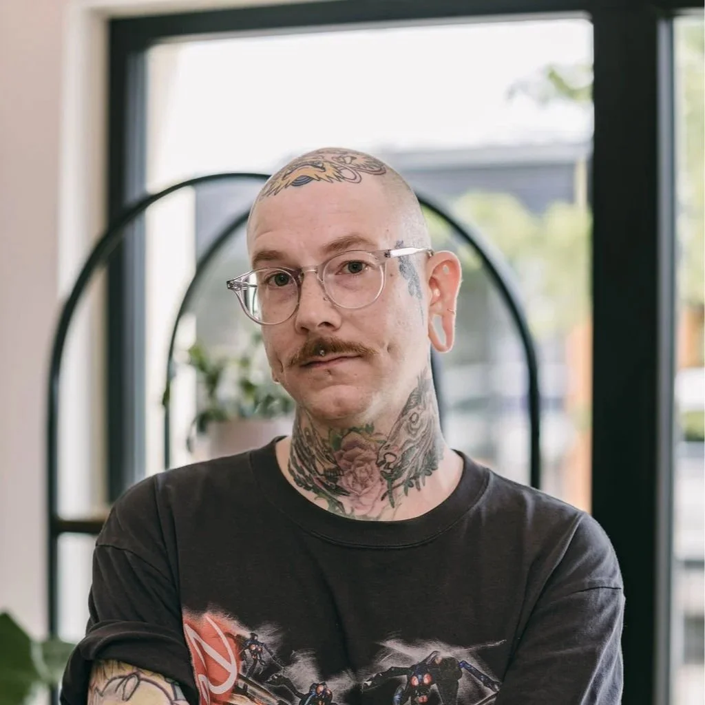 A tattooed man with glasses, a mustache, and piercings, wearing a black graphic T-shirt, standing indoors with a large window and plants in the background.