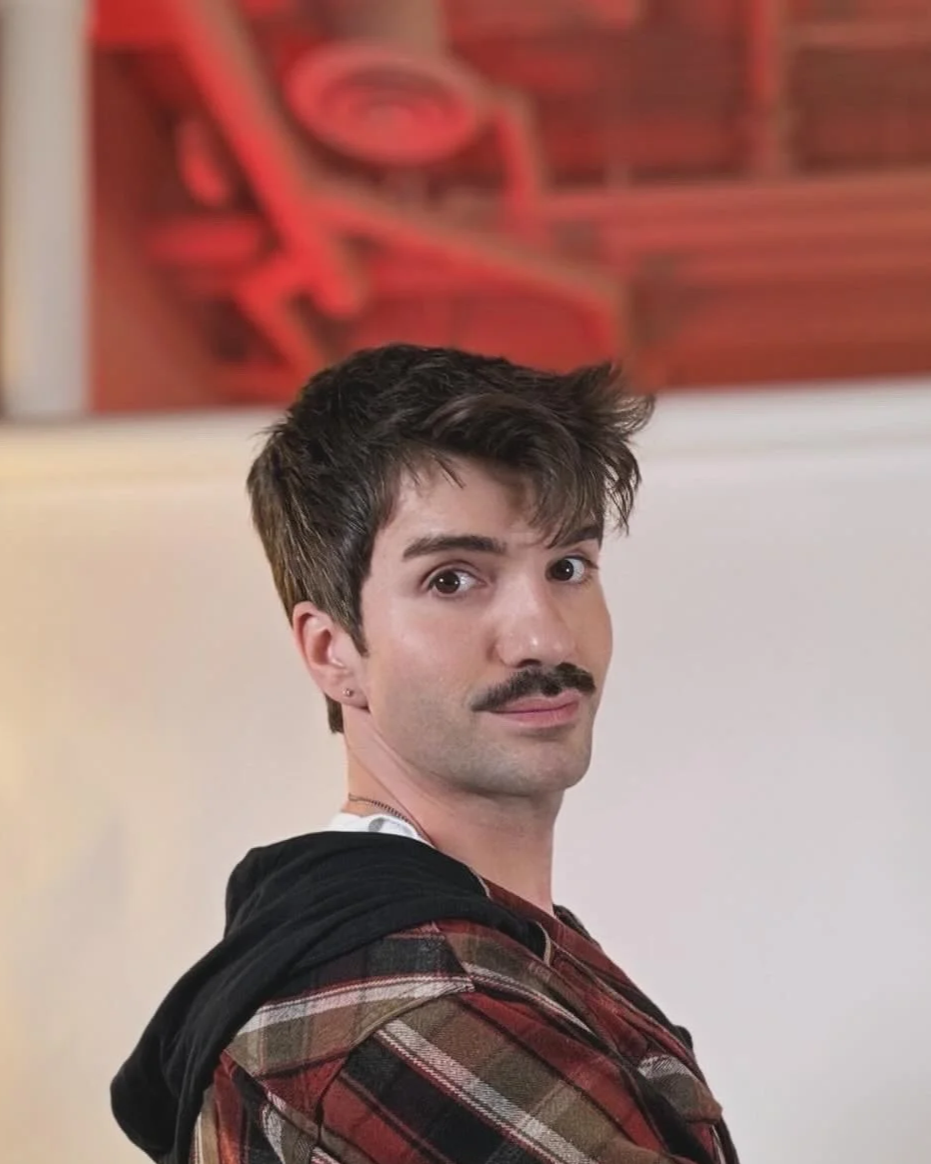 A young man with dark hair and a mustache looks over his shoulder, wearing a plaid shirt with a black hoodie draped over his shoulders, in front of a red piece of abstract art on a wall.