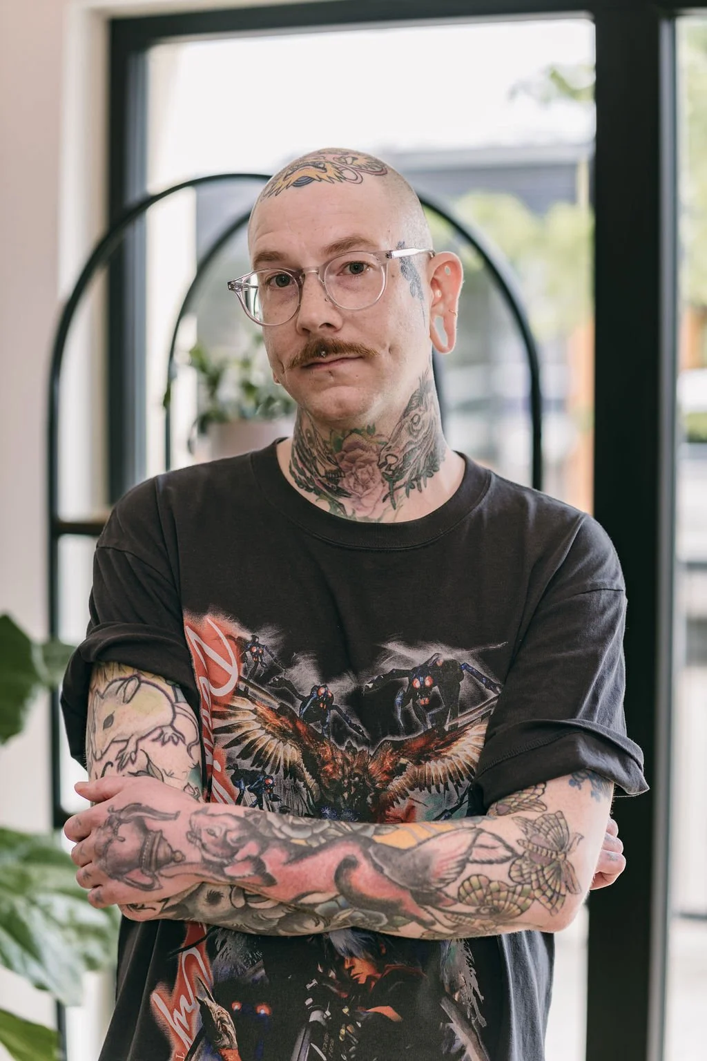 A tattooed man with glasses and a mustache posing indoors in front of a window, wearing a black graphic T-shirt with a sci-fi or fantasy design.