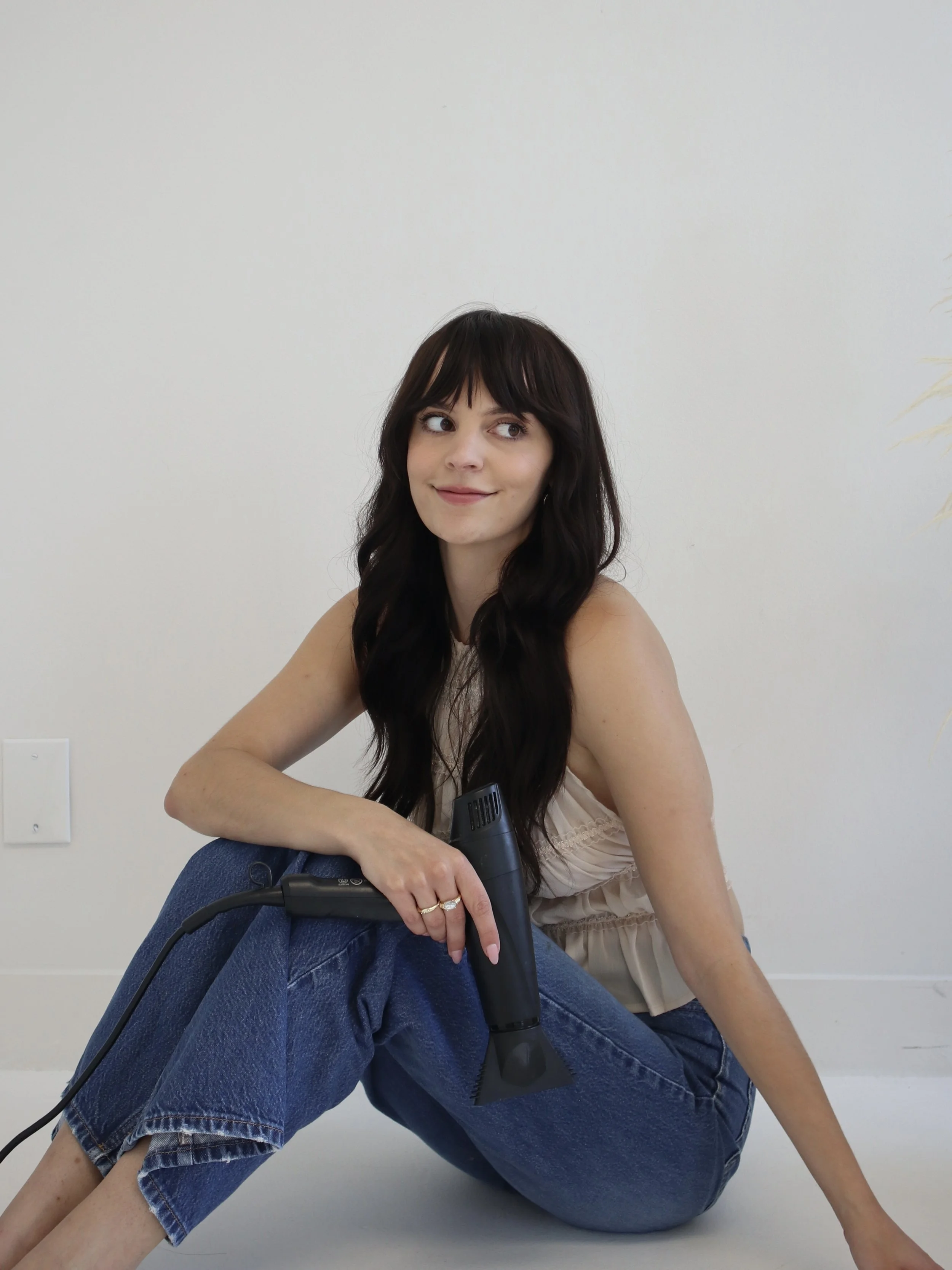 A woman with long dark hair and bangs sitting on the floor against a plain white wall, holding a black hair dryer, wearing a sleeveless top and blue jeans, with a slight smile and looking to the side.