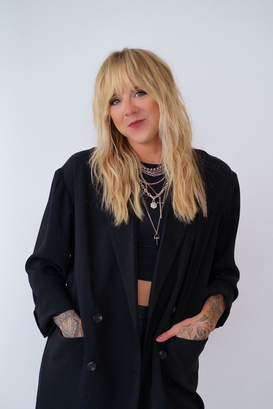 A woman with long blonde wavy hair and tattoos on her arms, dressed in black clothing with layered necklaces, standing against a plain light background.
