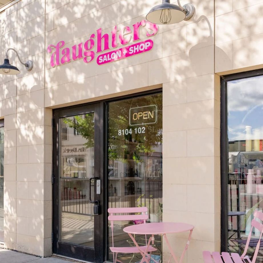 The exterior of a salon and shop called 'Laughters,' with a pink sign, a glass door with an 'OPEN' sign, and pink outdoor furniture.