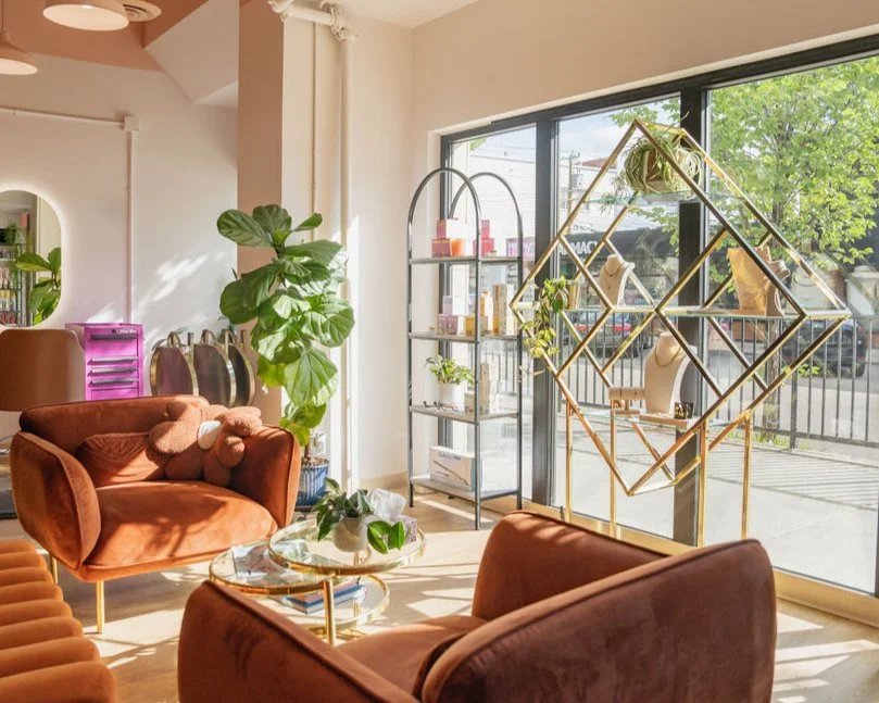 Interior of a vibey salon with warm brown armchairs in a cozy lounge,  green plants, a glass coffee table, gorgeous retail space with accessories candles and hair products, and large glass windows showing the busy street outside.