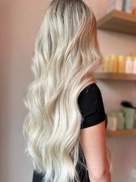 Side view of a woman with long, wavy, blonde hair with long hear extensions wearing a black shirt, standing indoors near shelves with colorful Design Me, Oribe, and R+Co Hair products.