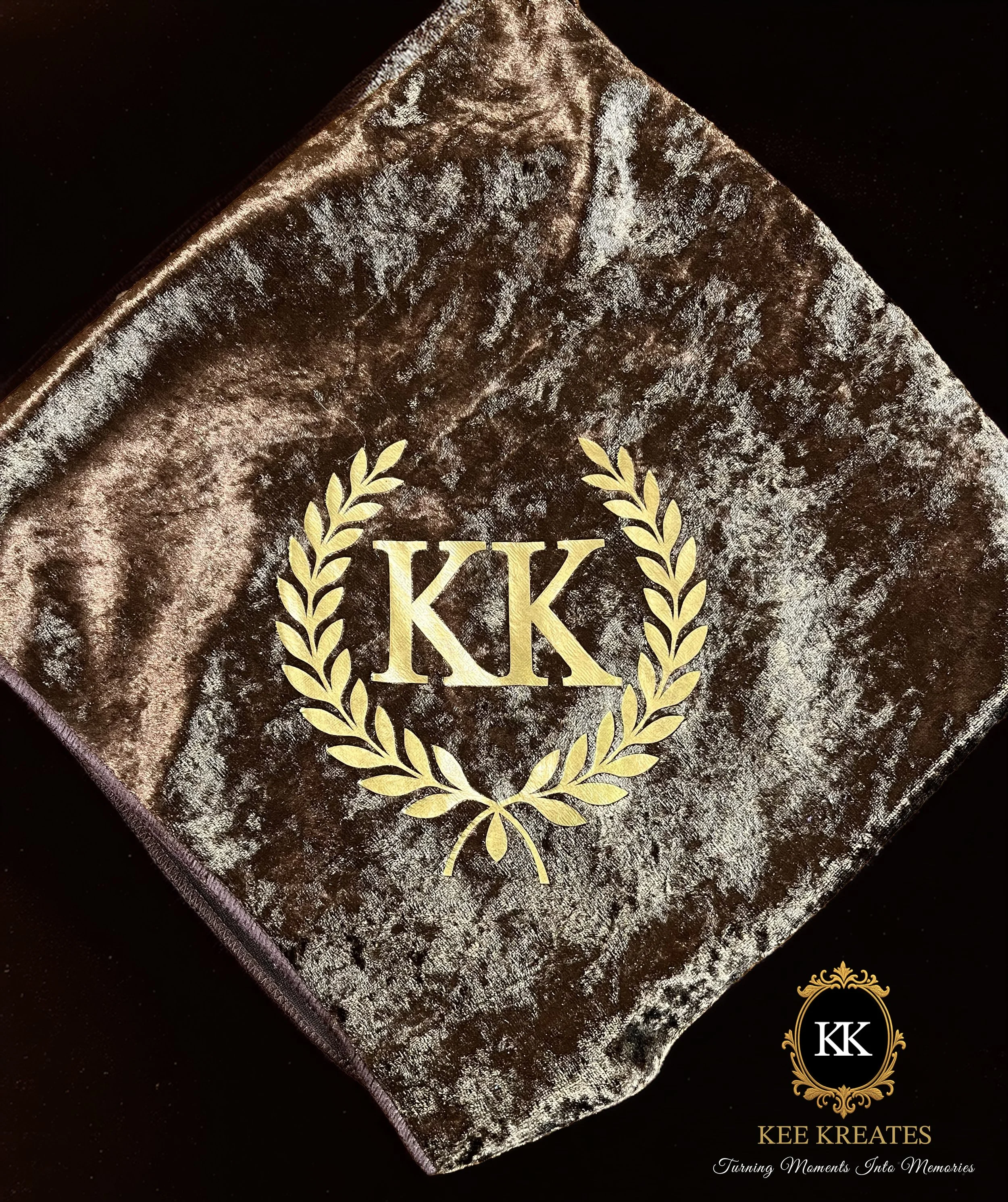 Kee Kreates Photo. A close-up of a velvet cloth with a large gold monogram 'KK' inside a gold laurel wreath, on a dark background. In the lower right corner, there is a logo with the initials 'KK' within an ornate gold frame and the text KEE KREATES.