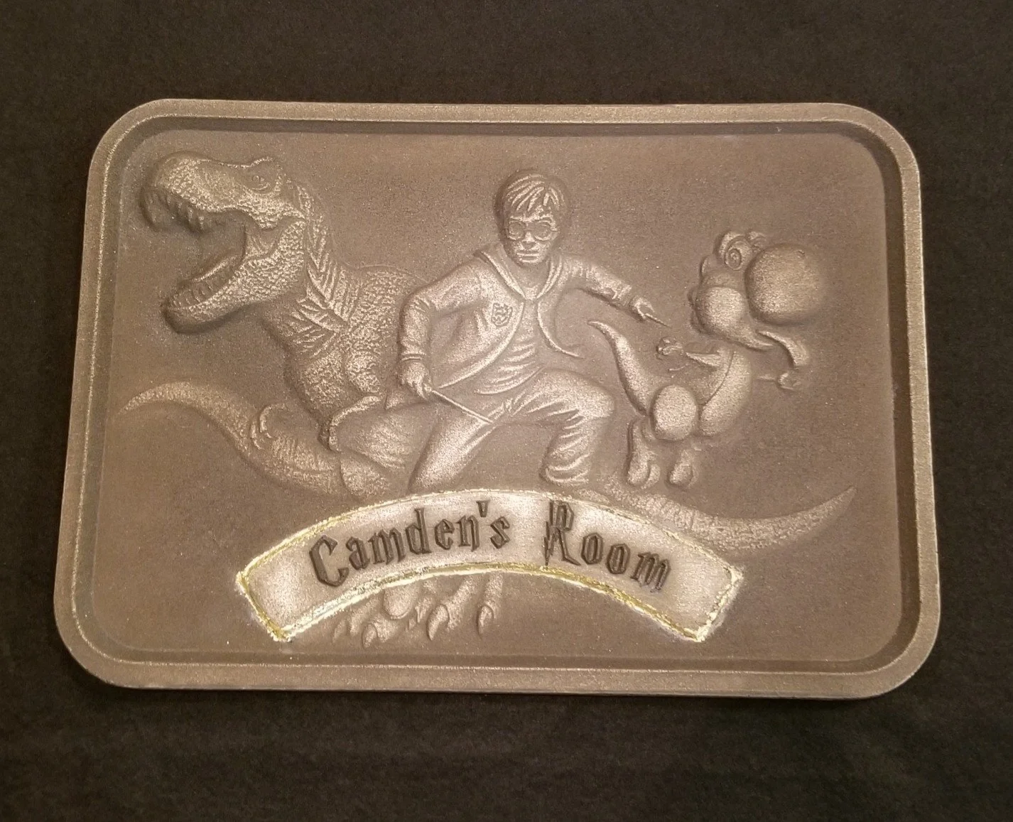 Bronze metal collectible plaque with embossed design of a dinosaur with a long tail and small arms and movie characters. The plaque has a banner at the bottom with the text "Camden's Room."