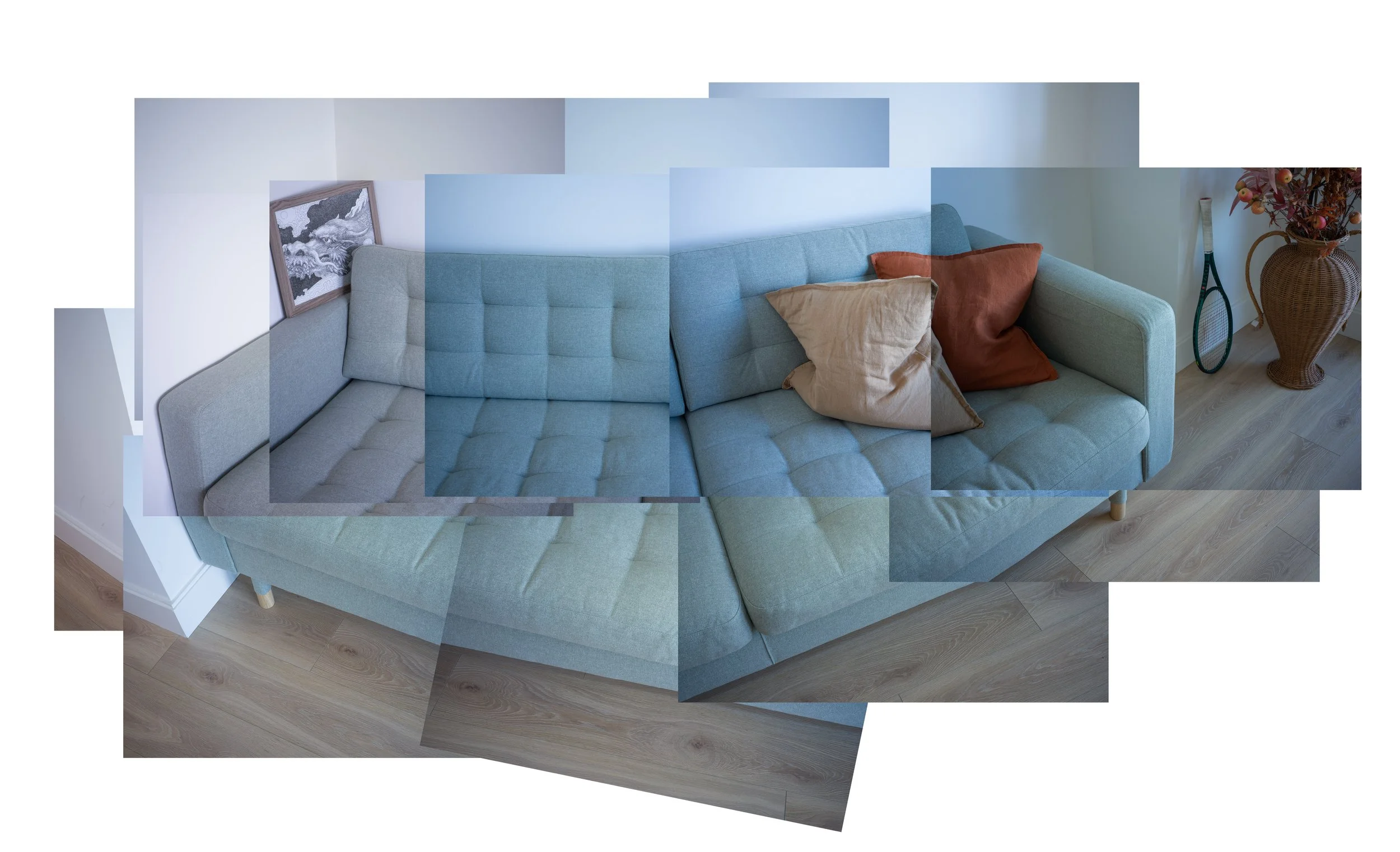 A light blue sectional sofa with several pillows on it, a framed artwork leaning against the wall, a tennis racket propped against a large rattan vase with flowers, and wooden flooring.