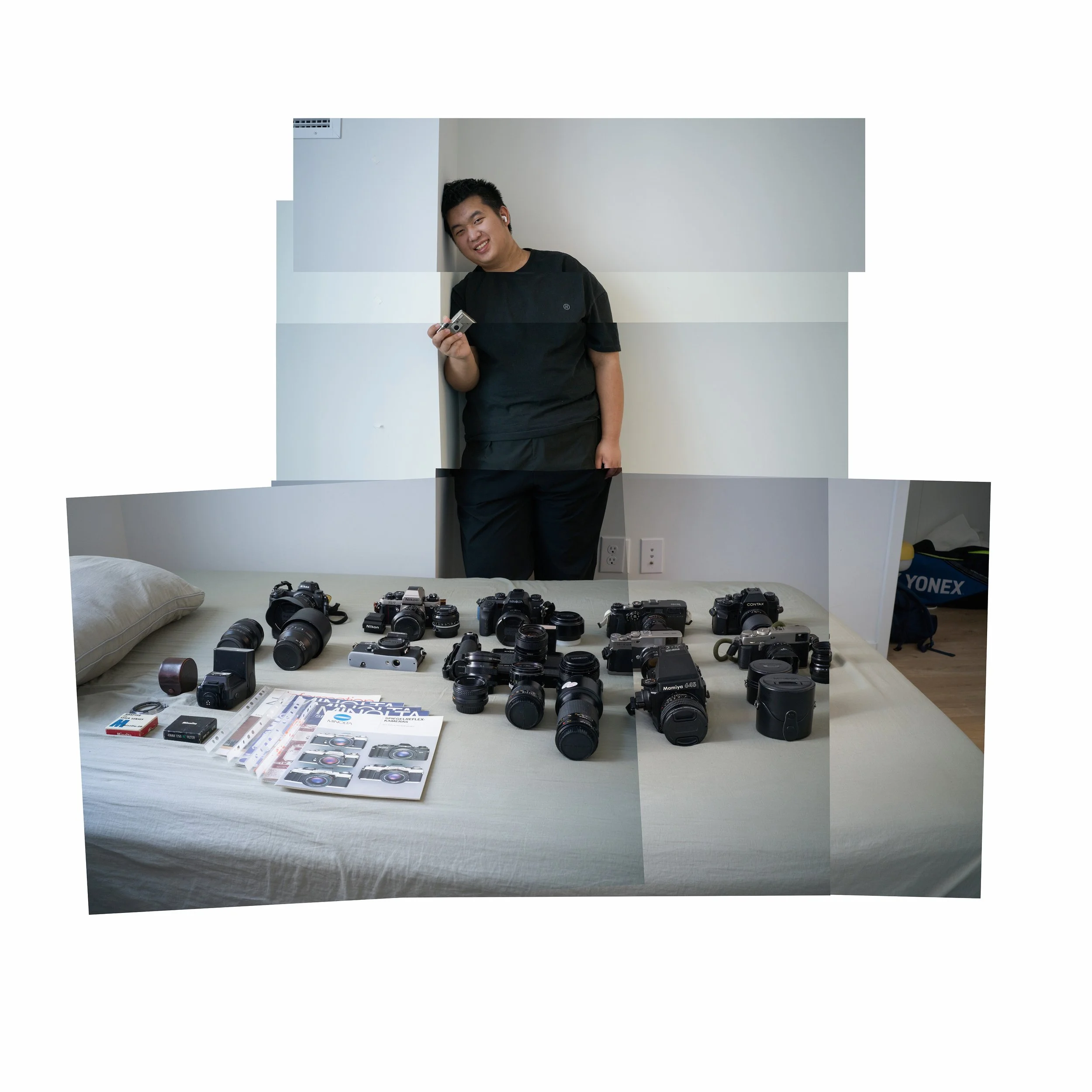 A young man stands next to a bed with various cameras and photography equipment laid out on it. He is holding a camera and smiling.