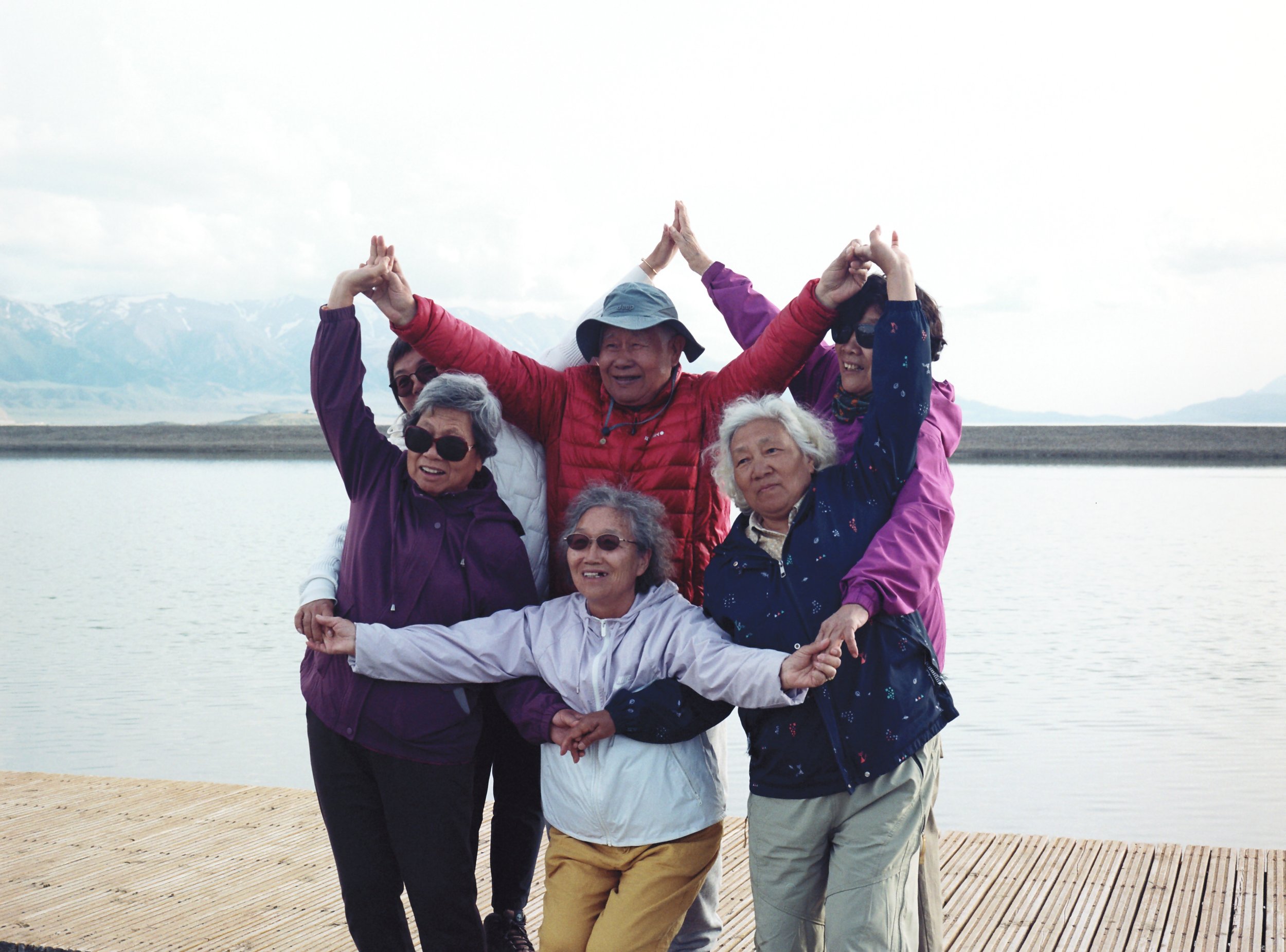 Seven elderly people, four women and three men, are posing together outdoors on a wooden deck by a body of water, with snow-capped mountains in the background. They are smiling and making playful hand and arm gestures, dressed in colorful jackets and