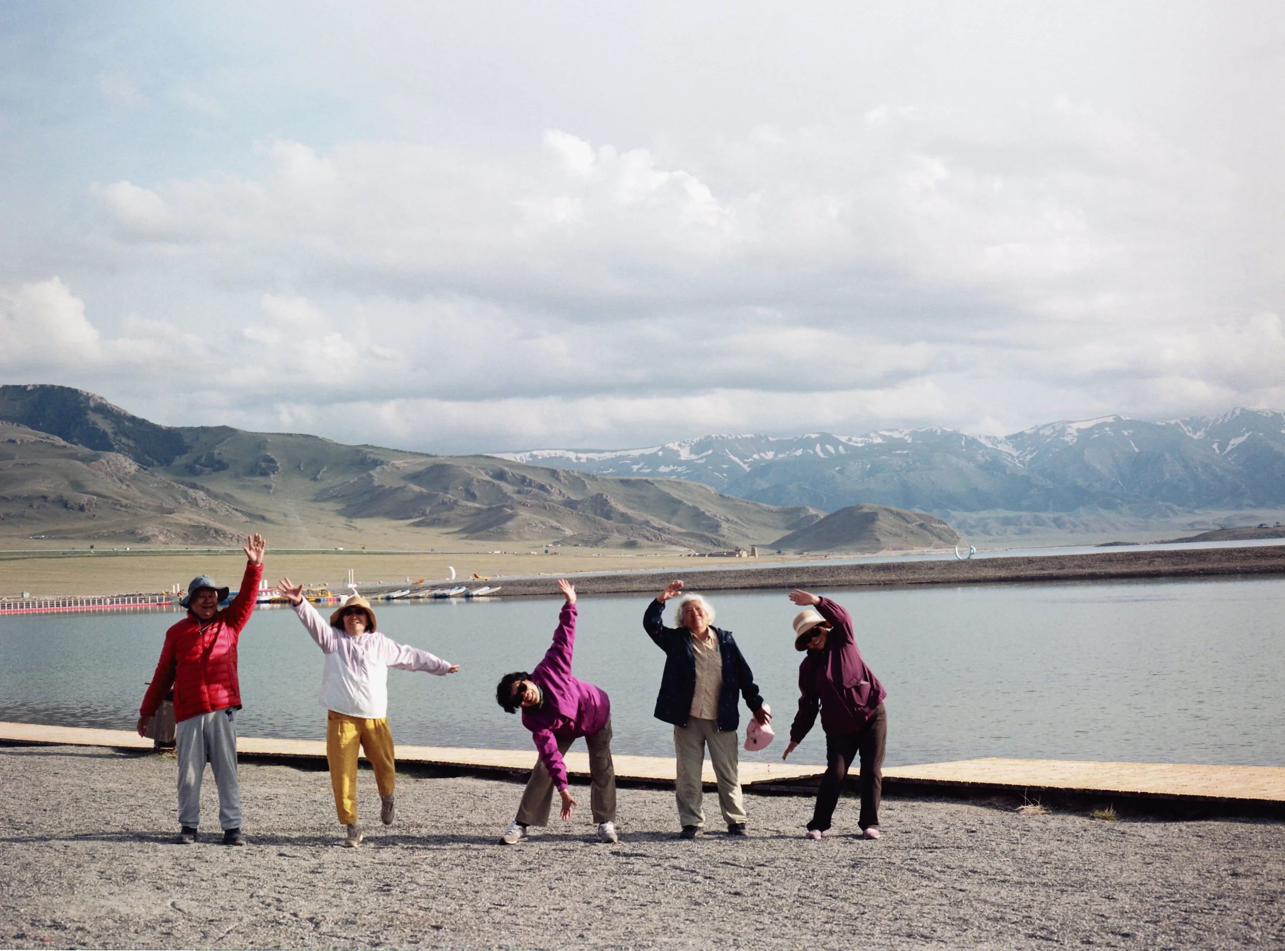 Five women standing by a lake with mountains and cloudy sky in the background, posing with playful and joyful gestures.
