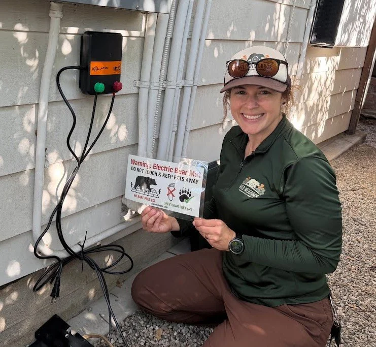 Woman in outdoor gear holding a warning sign about electric bear fencing, next to electrical equipment on the side of a house.