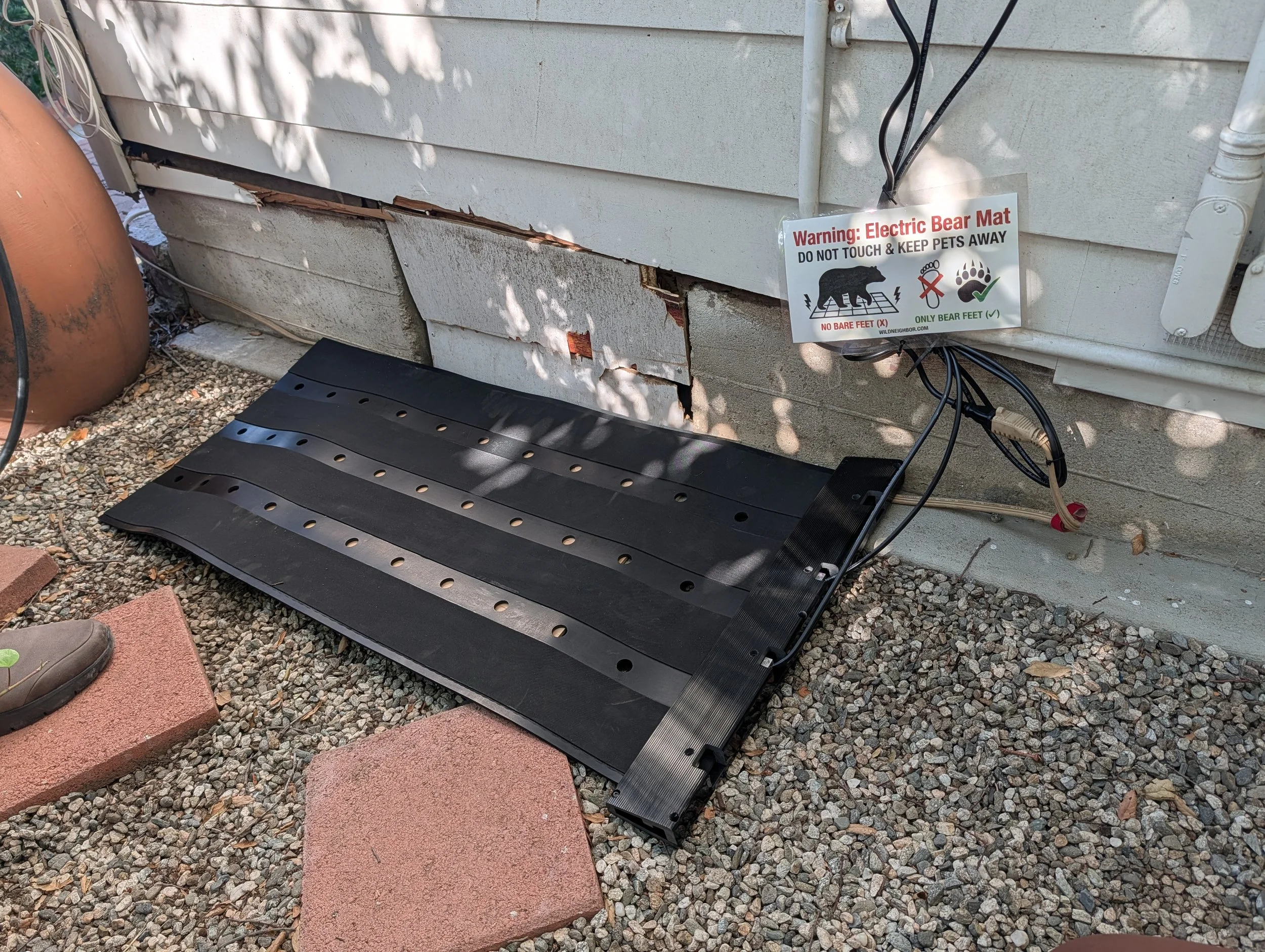 The image shows a damaged house exterior with a sign warning about an electric bear mat. The mat is placed on the ground near the house foundation, which has visible damage with peeling paint and wood rot. The warning sign indicates the mat is for bears and instructs not to touch or have pet contact with the device.