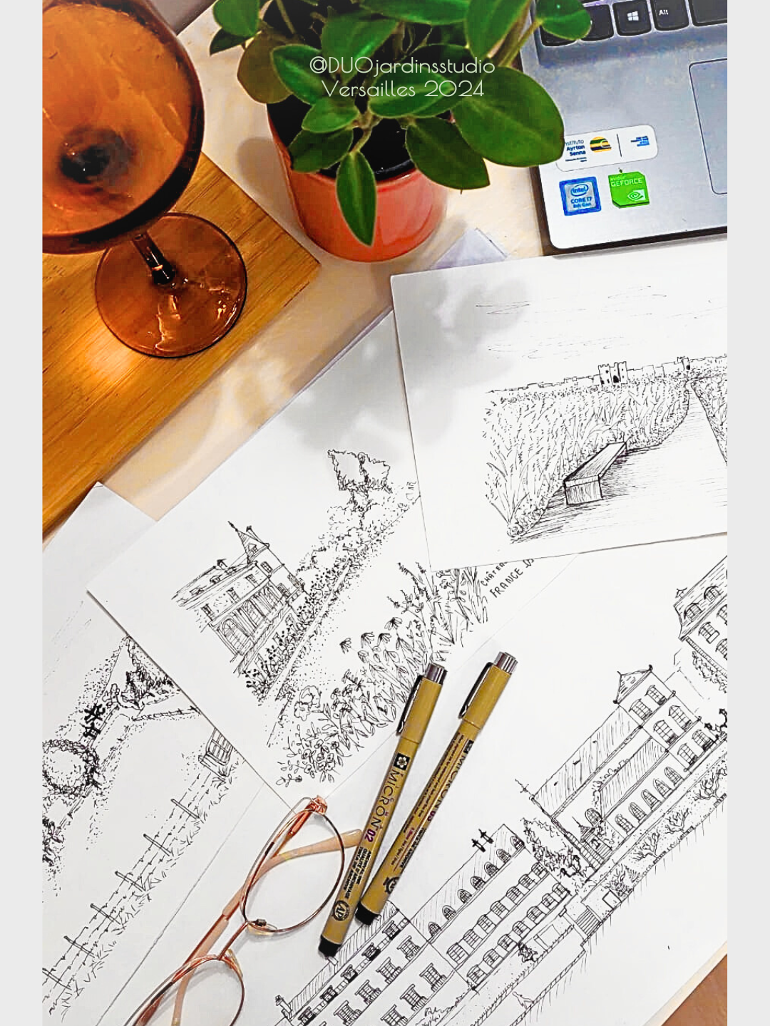 Croquis de paysages et bâtiments, deux stylos, une paire de lunettes, un verre de boisson rosée, une plante en pot et un ordinateur portable