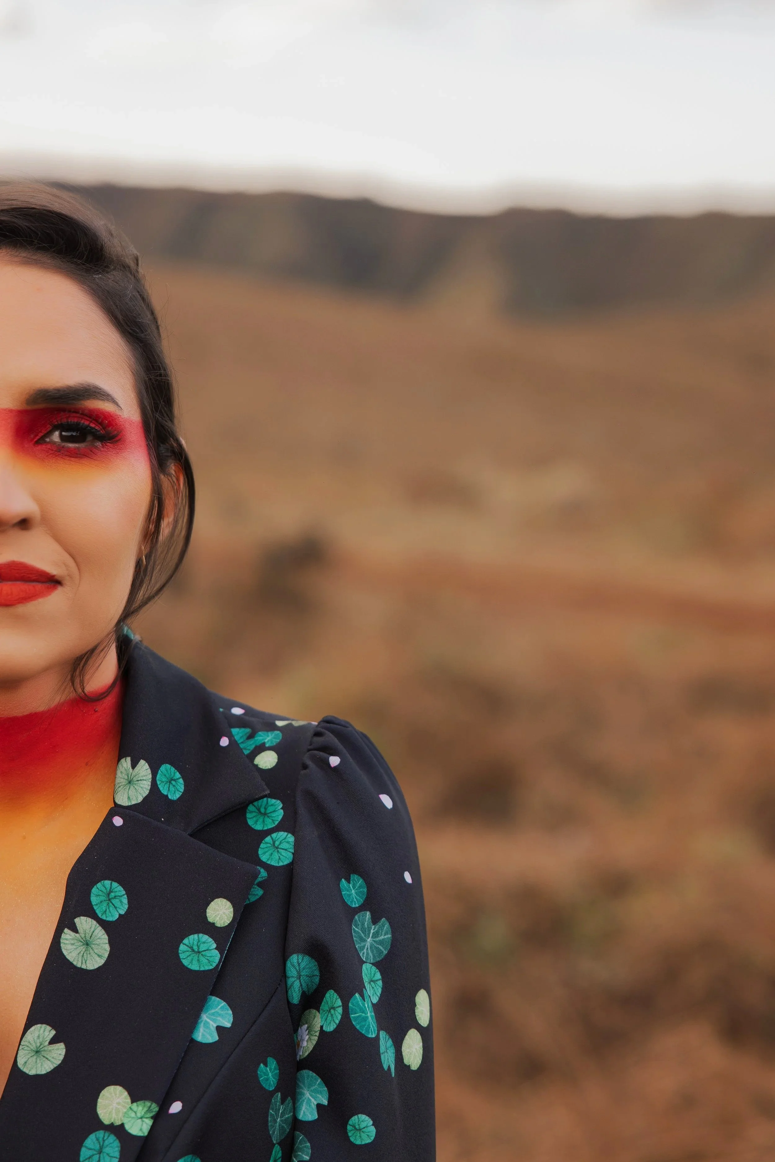 Une femme avec un maquillage coloré, portant une veste à motif de feuilles, dans un paysage naturel avec un ciel nuageux en arrière-plan.