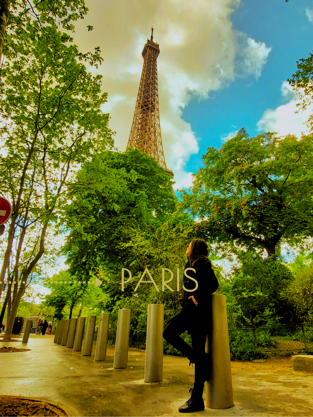 Une femme se tenant près d'une rangée de poteaux dans un parc verdoyant avec la Tour Eiffel en arrière-plan à Paris, sous un ciel partiellement nuageux.