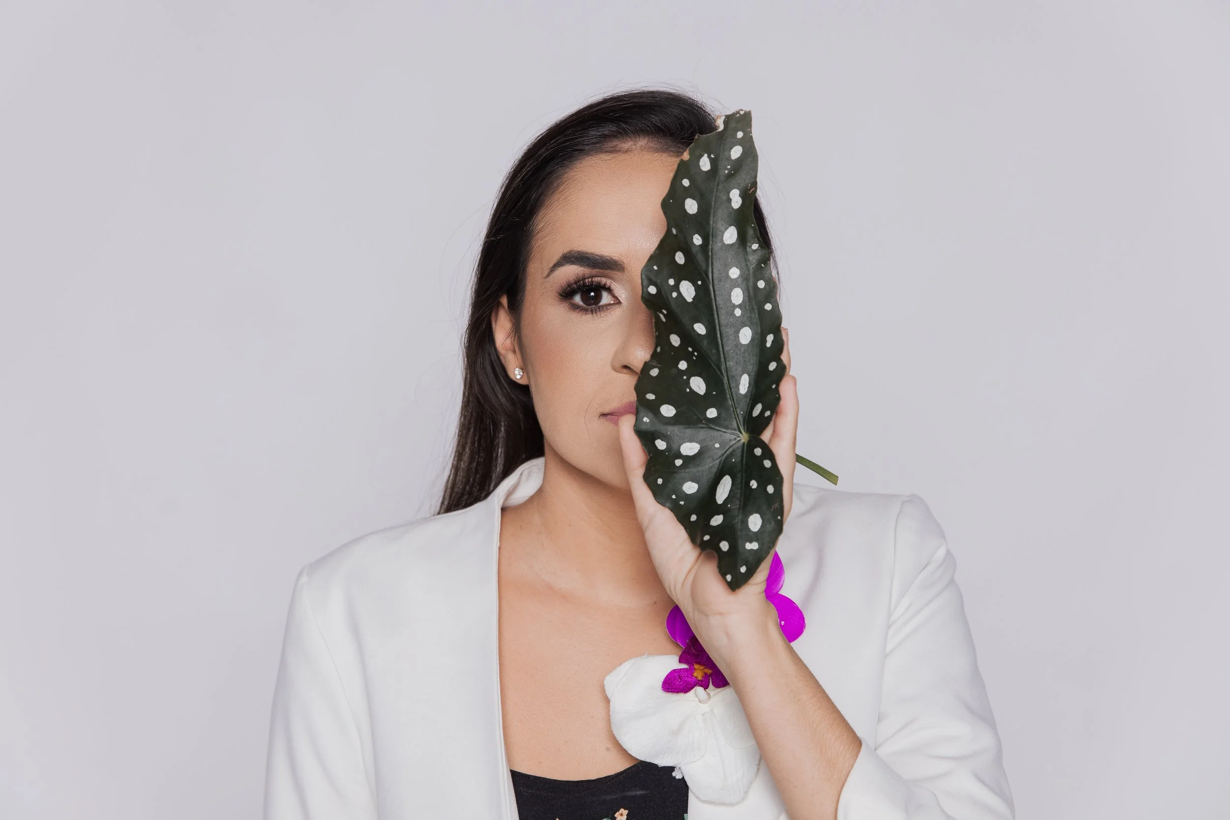 Femme avec un blazer blanc, tient une grande feuille vert foncé avec des taches blanches devant son visage, dans un fond gris clair.