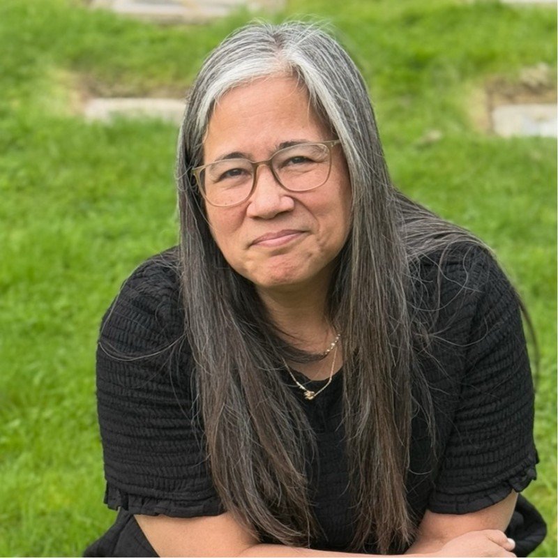 A woman with long gray hair and glasses sitting outdoors on a grassy area, wearing a black top and smiling slightly.