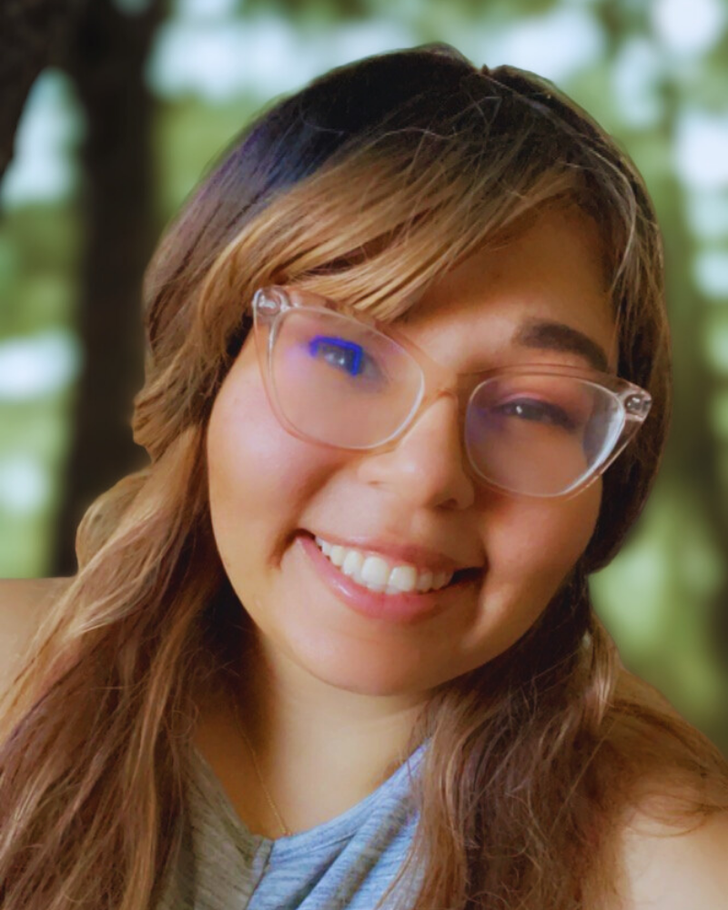Close-up of a young woman smiling, wearing glasses, with long wavy brown hair, outdoors in a green, forest-like background.