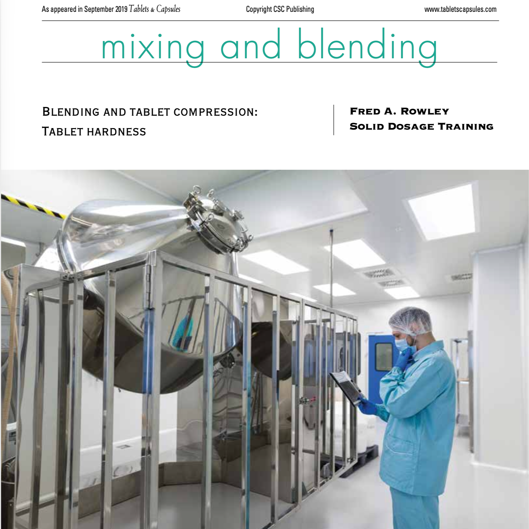 A person in a blue medical gown, mask, and gloves is standing in a laboratory, reading from a tablet device. In the background, there is a large stainless steel machine in a clean, well-lit room.