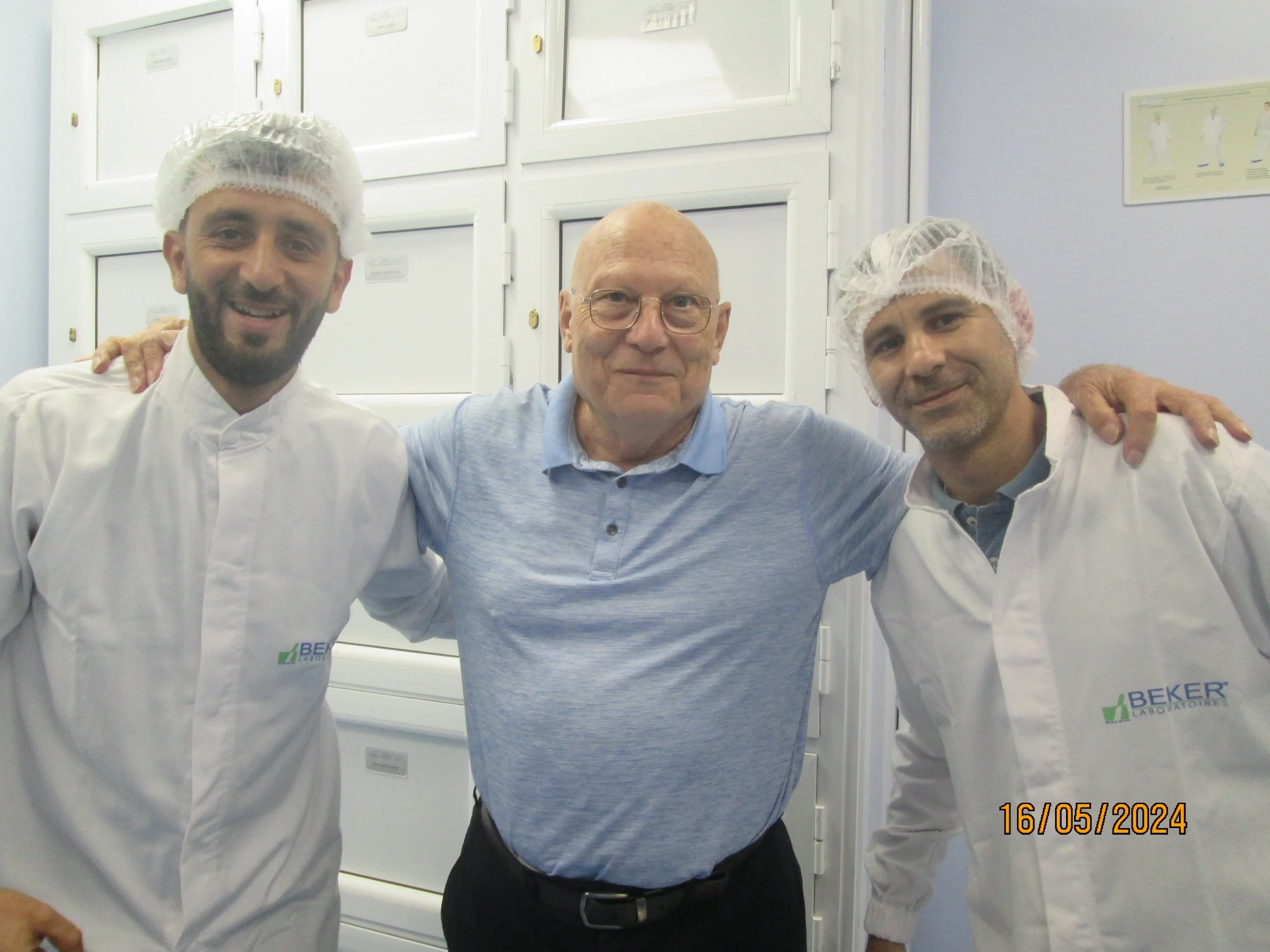 Three men smiling with arms around each other, two wearing white lab coats and hairnets, the middle man in a blue polo shirt, standing in front of white cabinets.