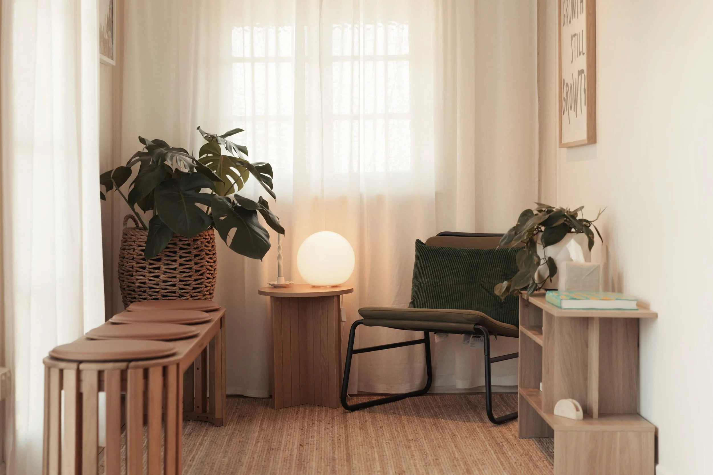 A cozy corner in a room featuring a wooden bench with a rounded top, a potted plant in a woven basket, a small round table with a spherical lamp, a green armchair with a dark cushion, and a wooden bookshelf with books and decorations. Light-colored curtains cover the window, allowing natural light to brighten the space.