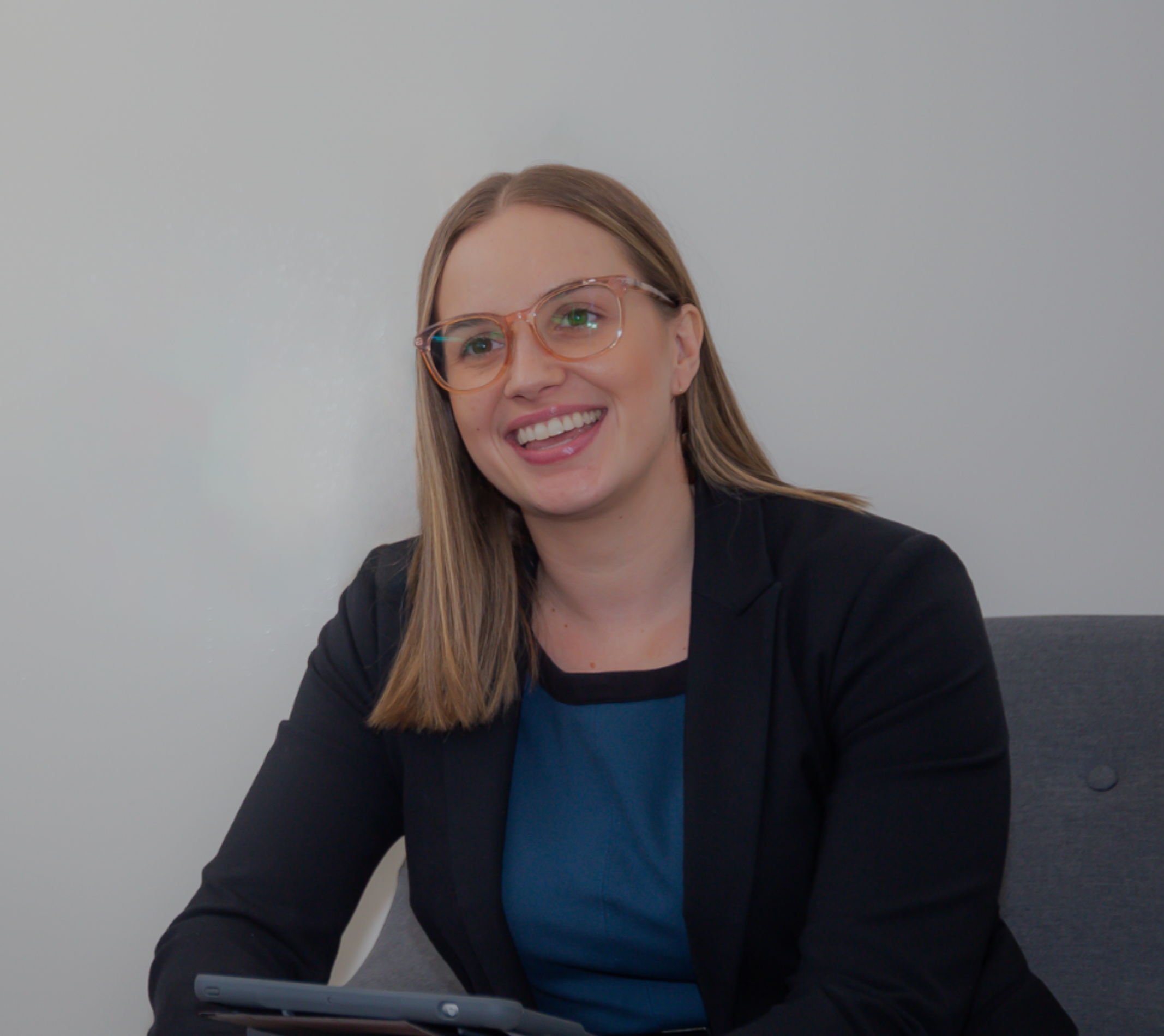 A young woman with light brown hair and glasses smiling while looking at her tablet. She is dressed in a dark blazer and a blue top, sitting against a plain light-colored background.